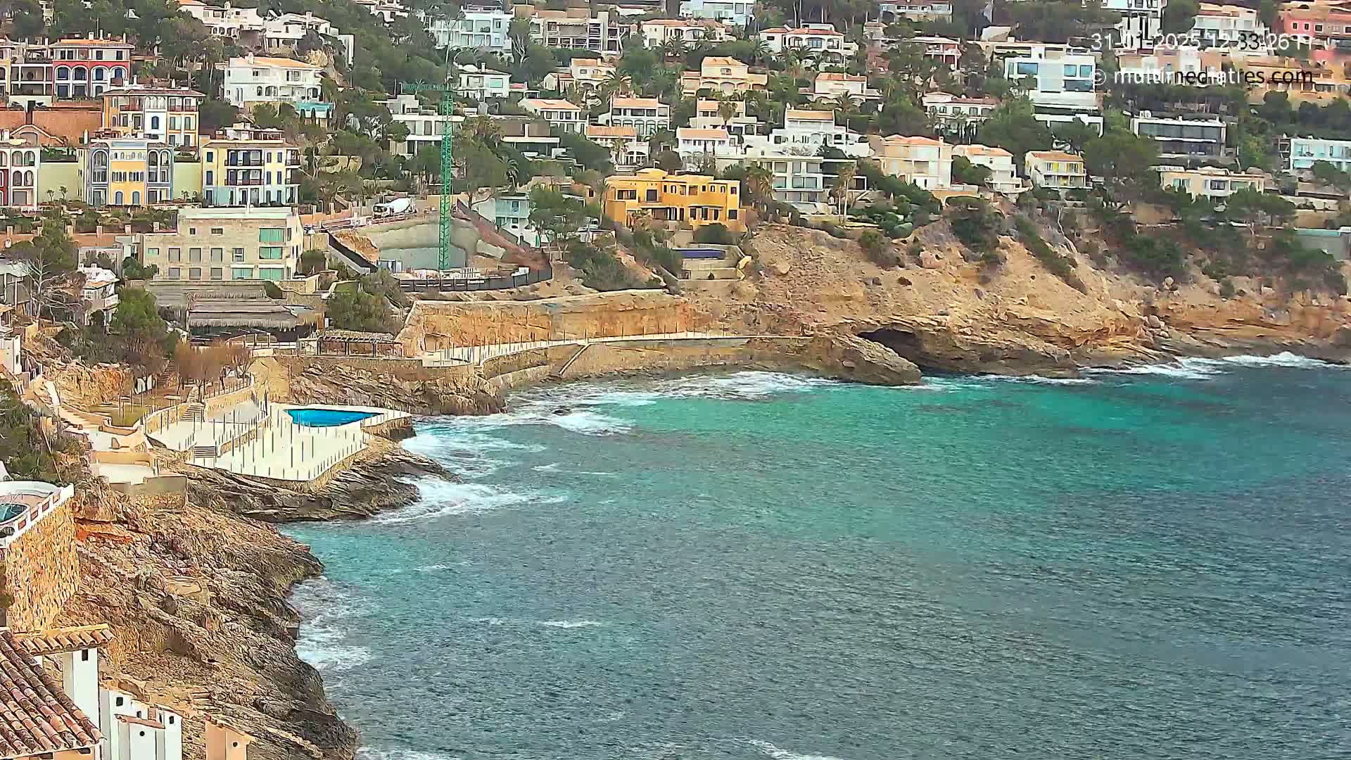 A rocky coastline with turquoise water features numerous houses built on a hillside, a small swimming pool, and some light wave action.