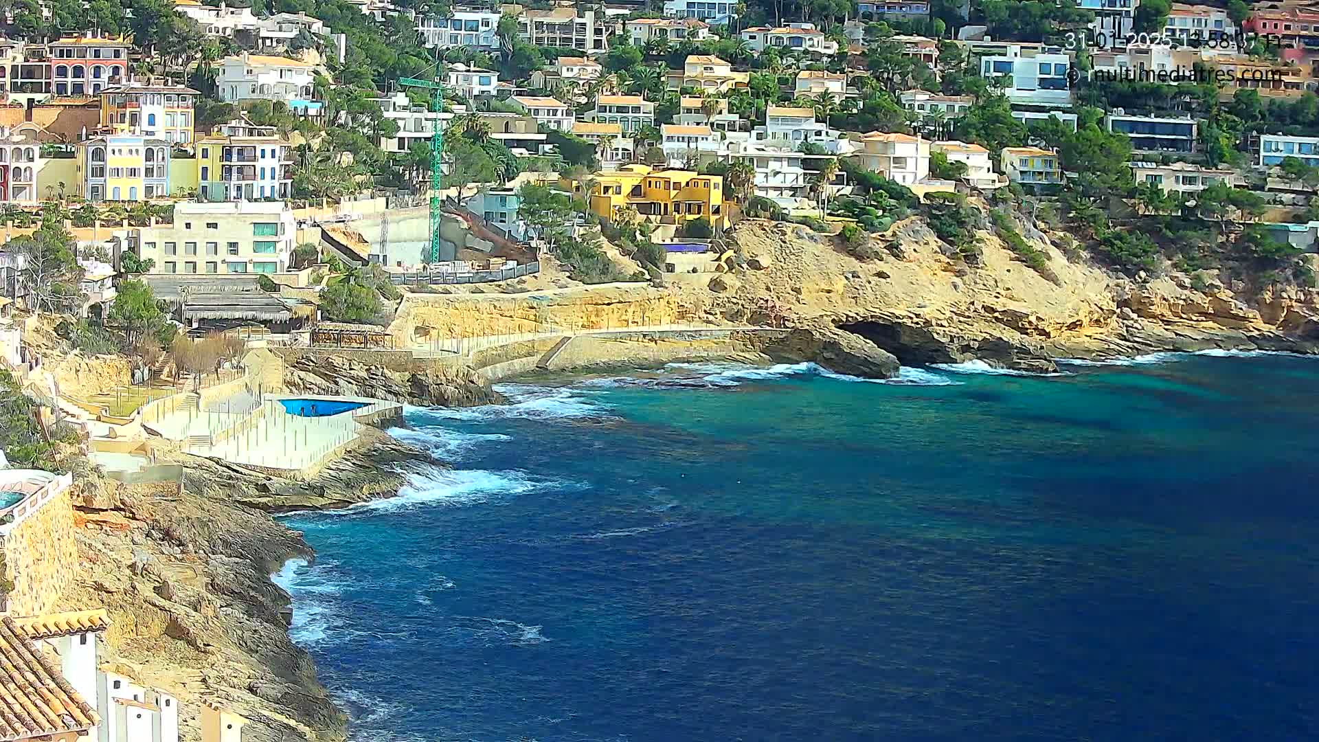A rocky coastline with a cluster of houses perched on the hillside overlooks a calm, teal ocean with small waves lapping against the shore.