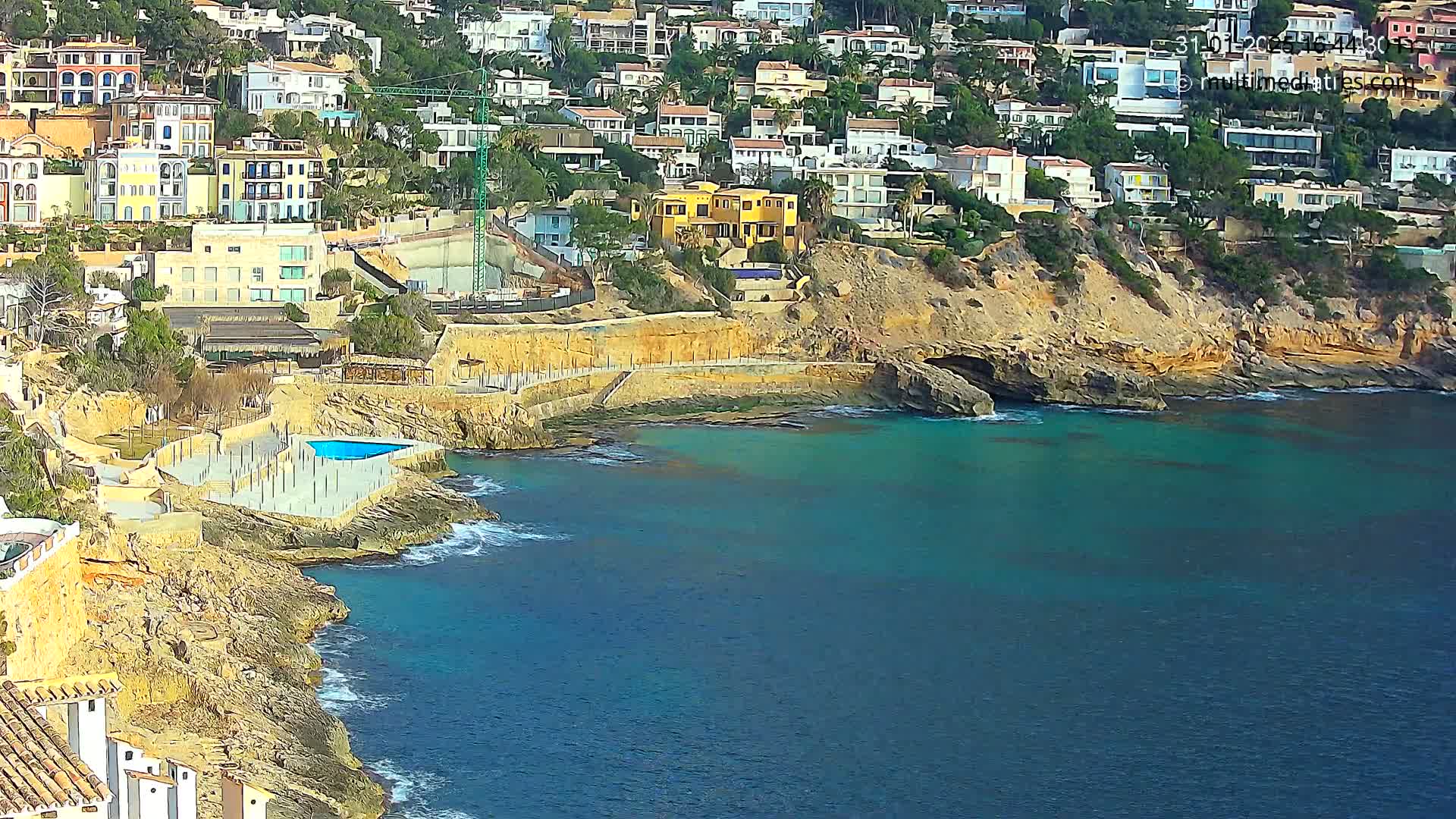 A rocky coastline with turquoise water features numerous houses built into a hillside, a small pool, and some construction.