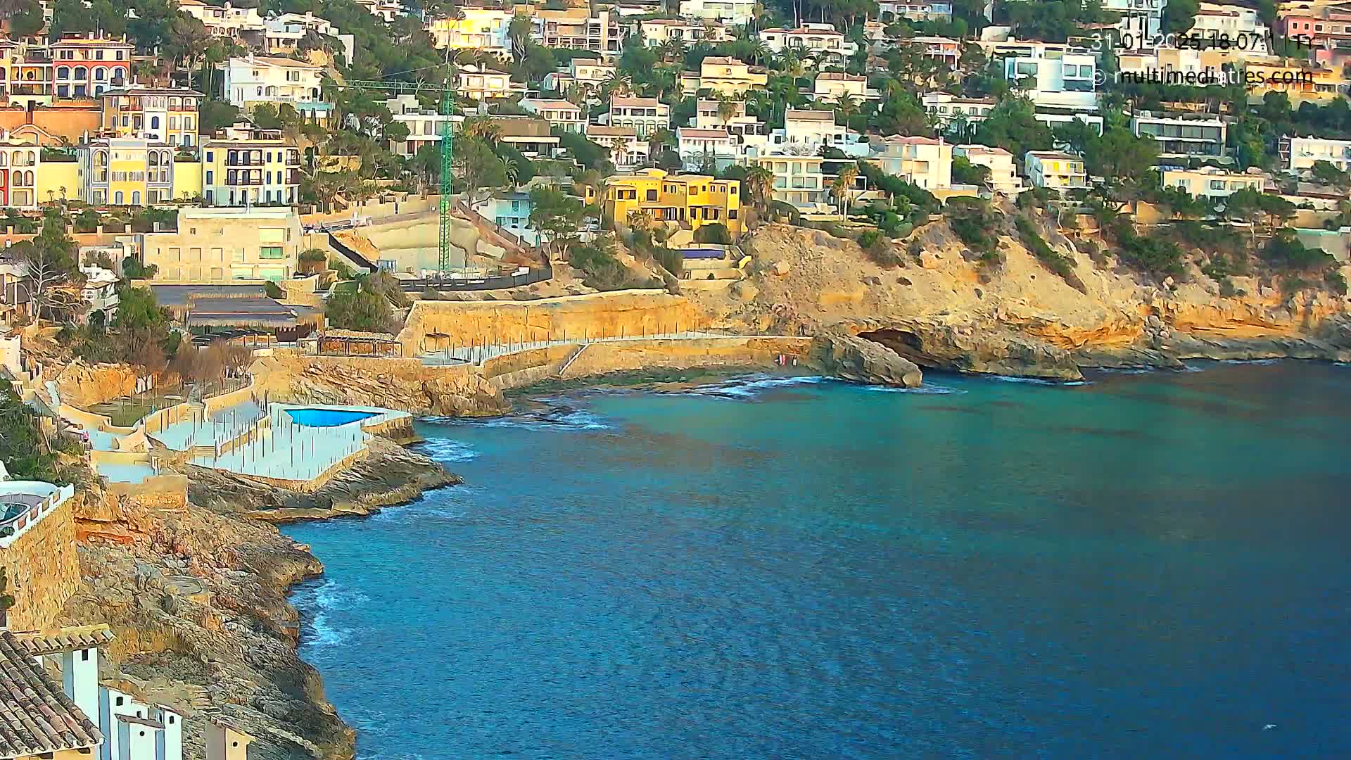 A coastal scene shows a hillside of multi-story houses overlooking a rocky cove with a turquoise sea.