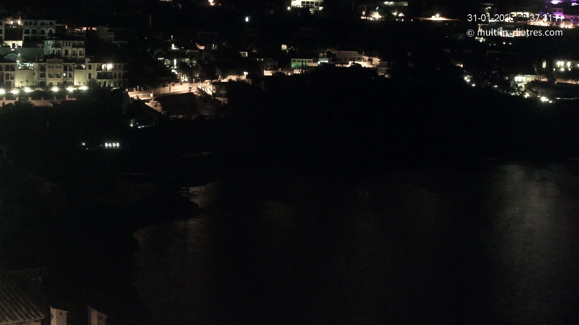 A nighttime, aerial view shows a coastal town with dimly lit buildings and a dark body of water.