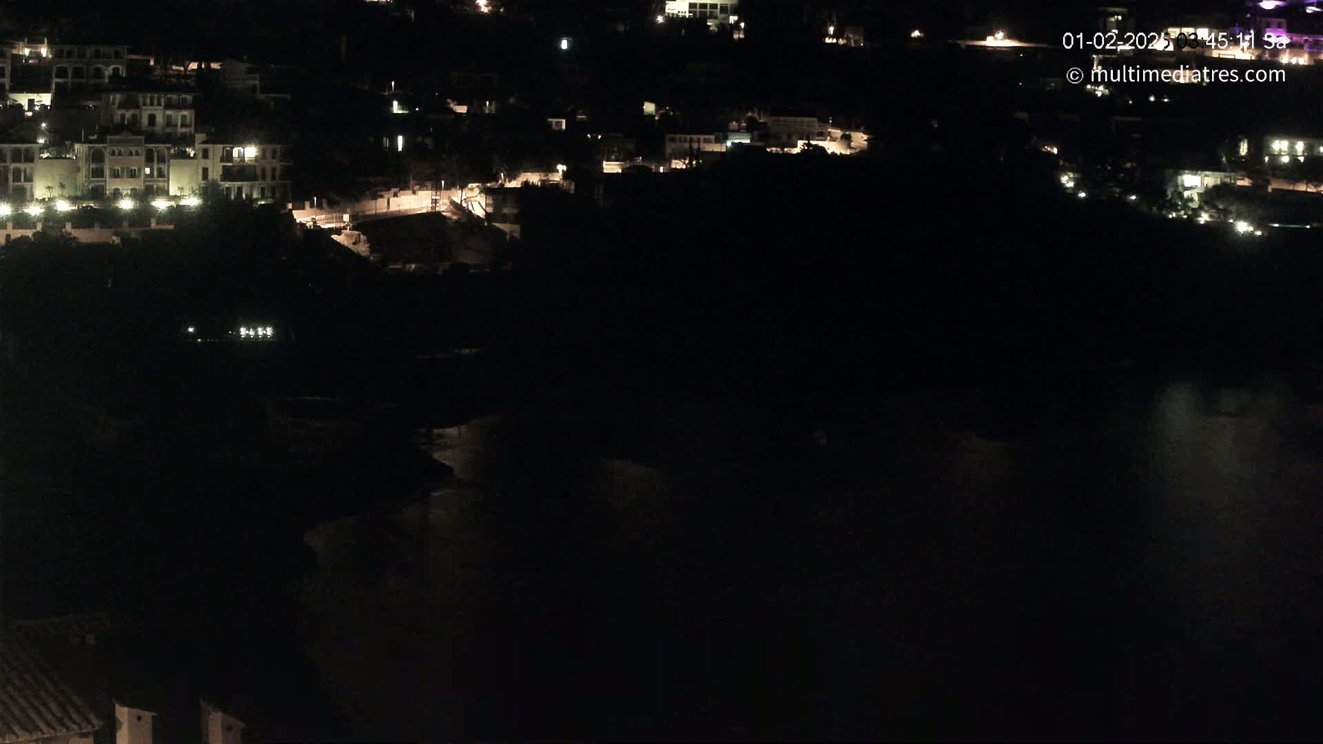 A nighttime view shows a dark body of water reflecting the lights of buildings on a hillside.