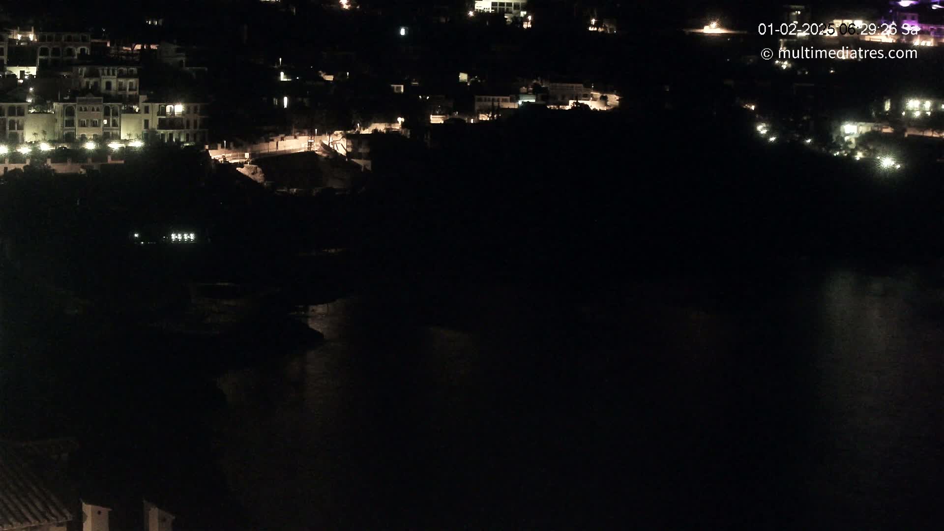 A nighttime, aerial view shows a dark body of water reflecting some light from a hillside town with sparsely lit buildings.