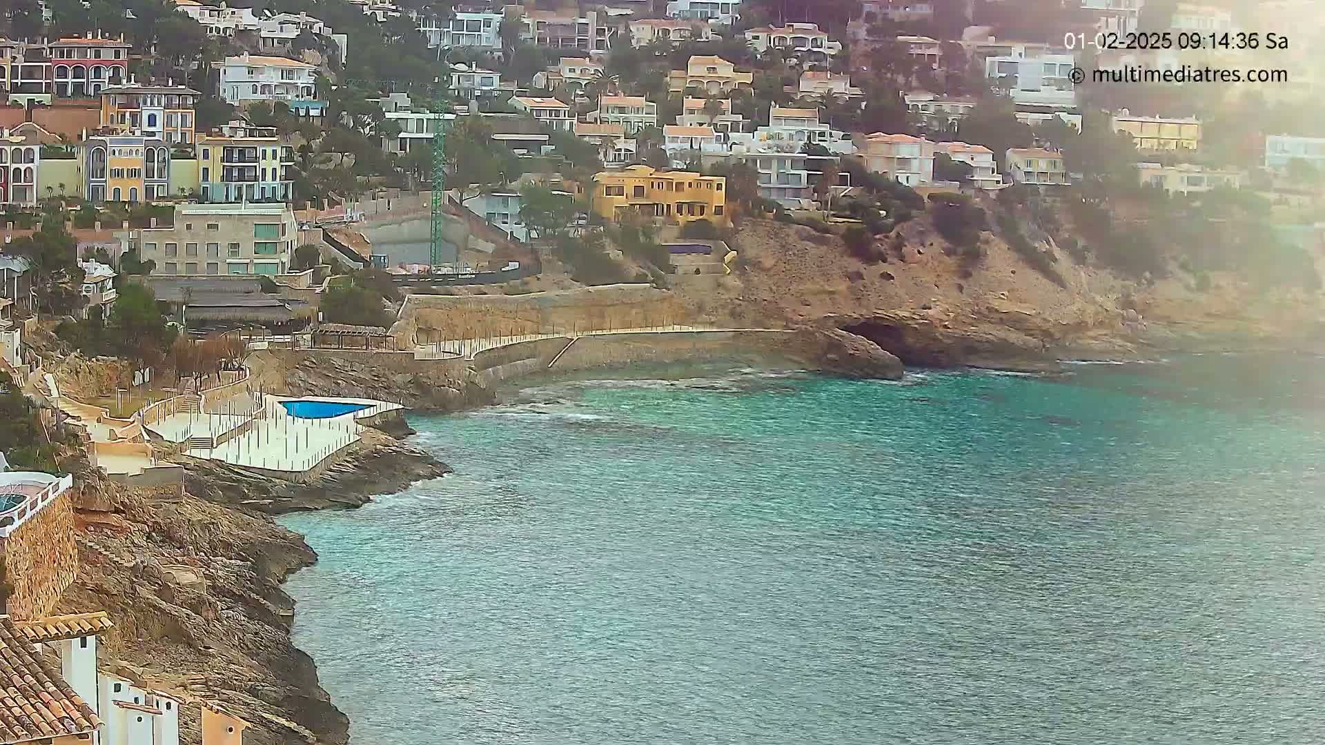 A rocky coast with a small, light-blue pool and many houses perched on a hillside overlooks a calm, turquoise sea.