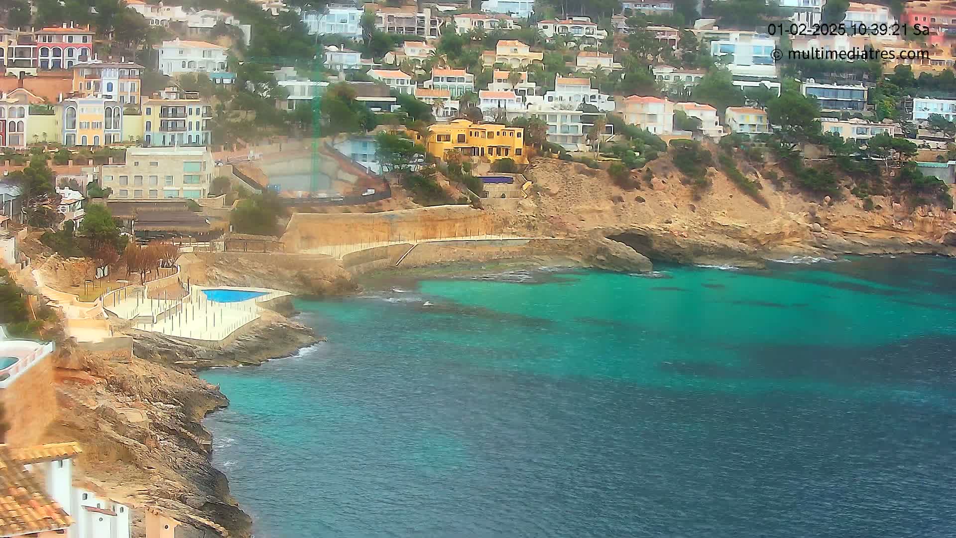 A rocky coastline with turquoise water features a small cove containing a pool and many houses built into the hillside.