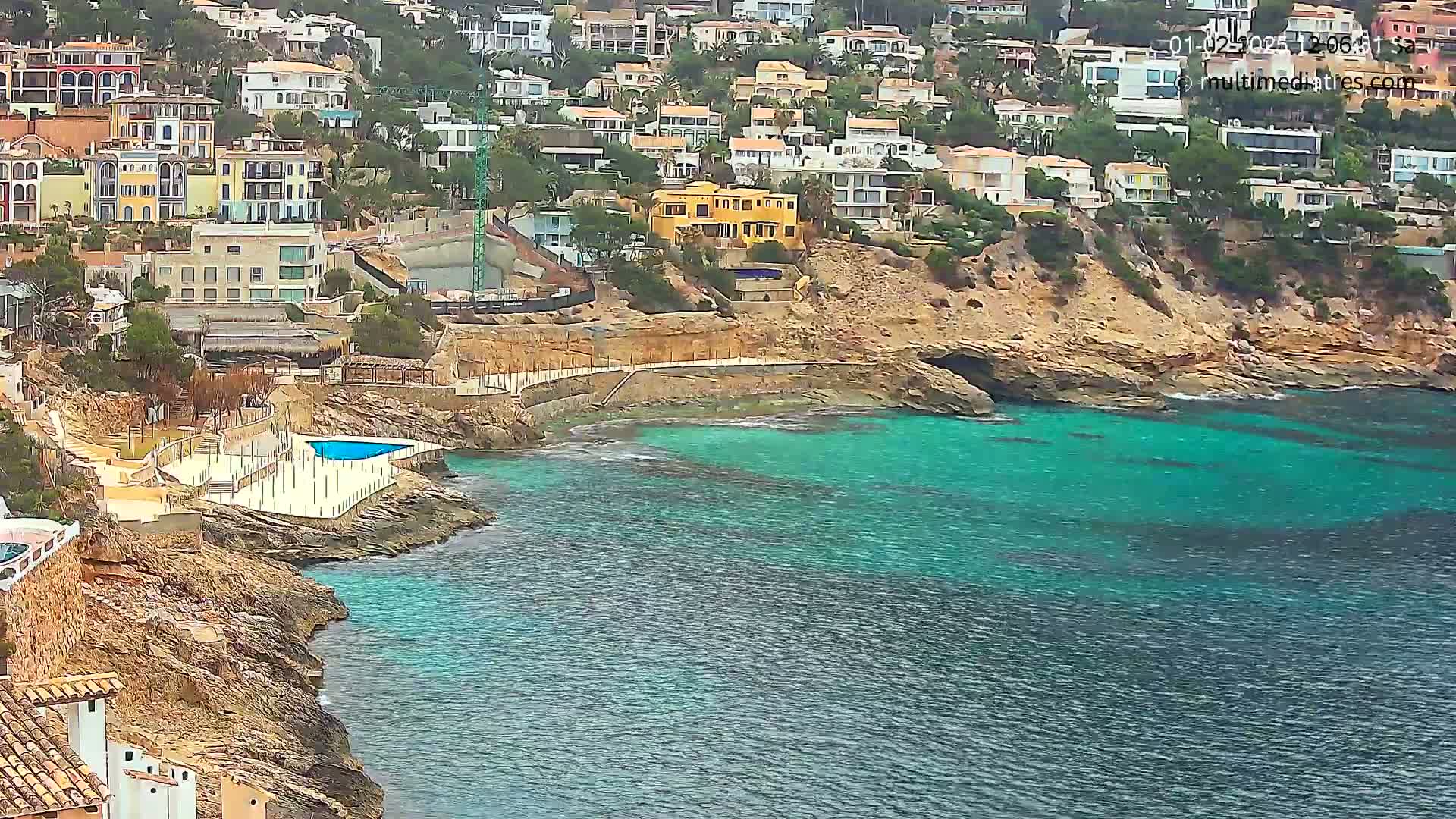 A rocky coastline with colorful houses perched on a hillside overlooking a calm, teal sea with a small pool visible near the shore.