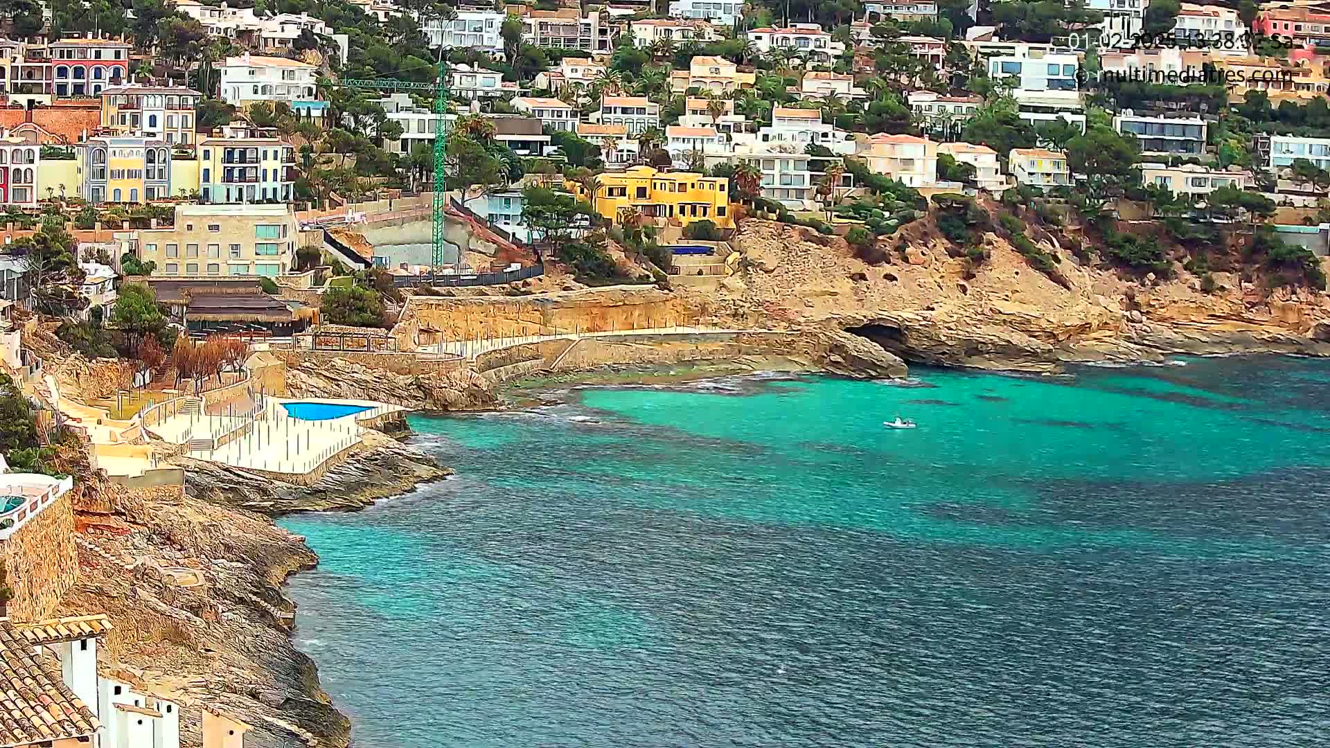 A coastal town with colorful houses built on a rocky hillside overlooks a calm, turquoise sea where a small boat is visible.