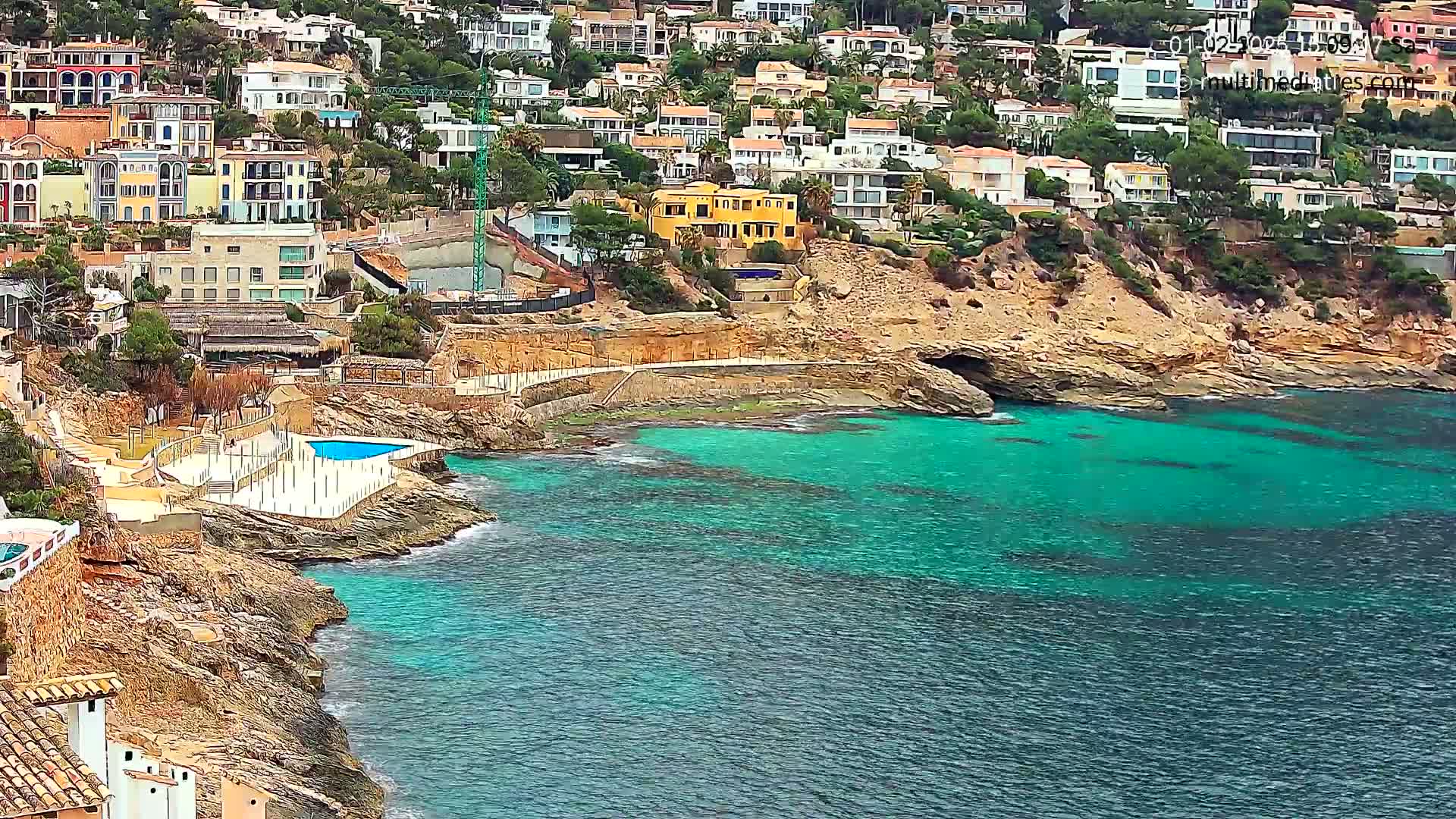 A coastal scene shows a hillside of multi-colored houses overlooking a teal-colored sea with rocky shores.