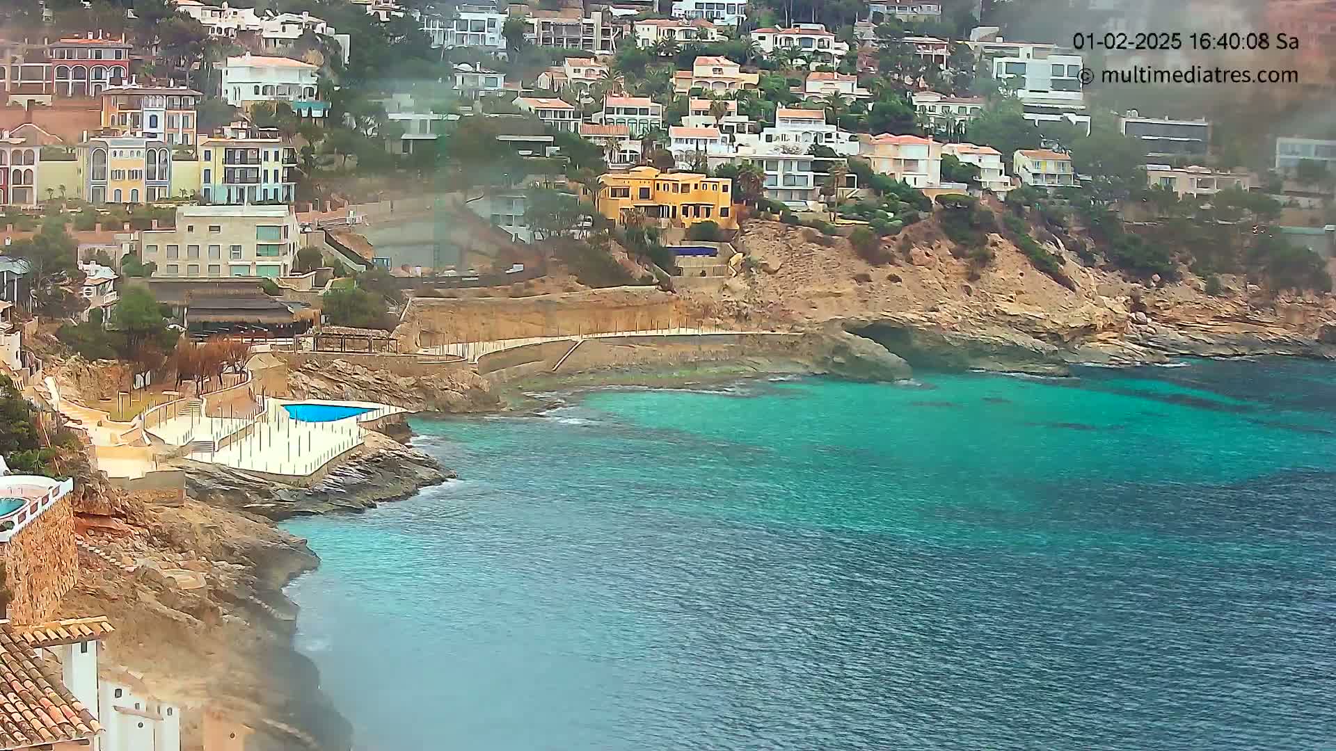 A coastal town with numerous houses built on a hillside overlooking a calm, turquoise sea on a slightly hazy day.