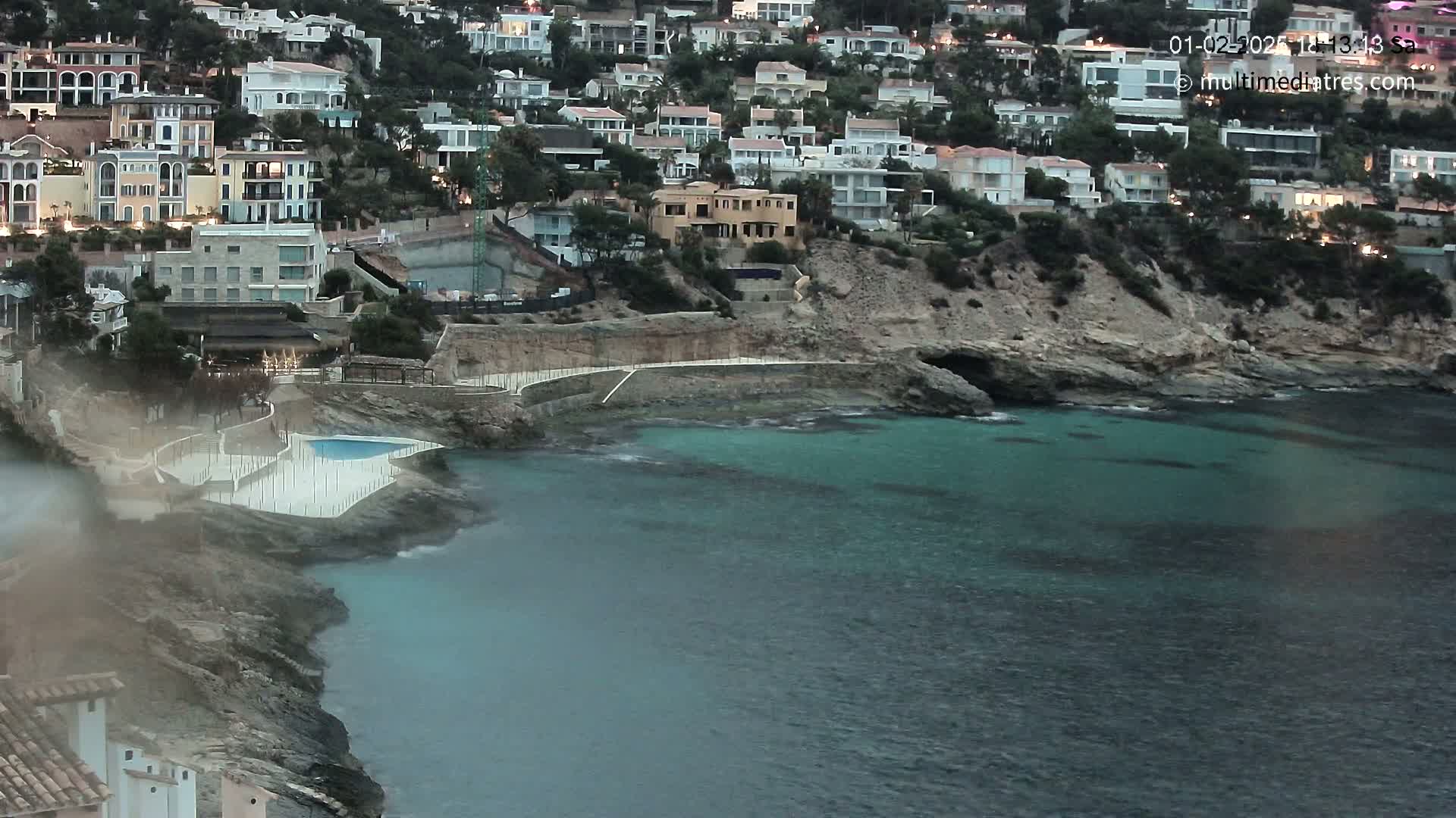 A coastal town with numerous houses sits on a rocky hillside overlooking a calm, teal sea with a small pool near the water's edge.