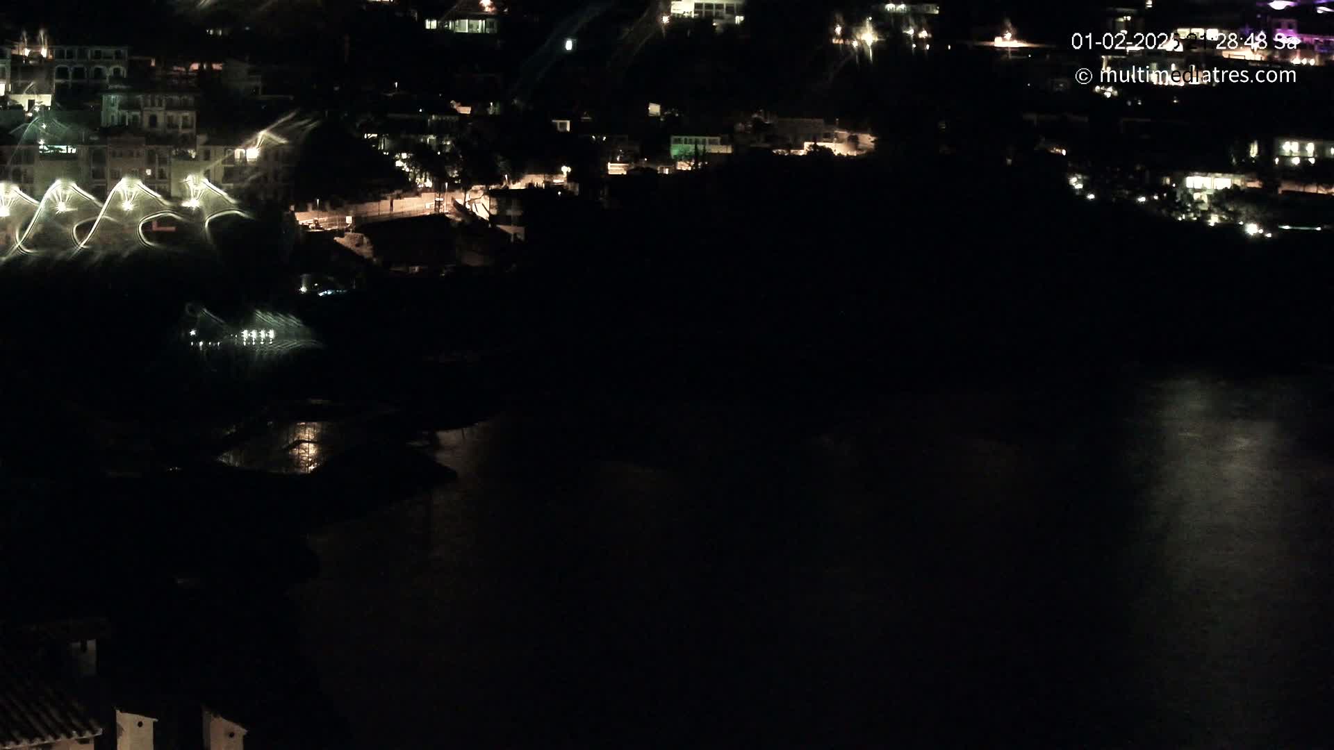 A nighttime, aerial view shows a dark body of water reflecting the lights of a hillside village.