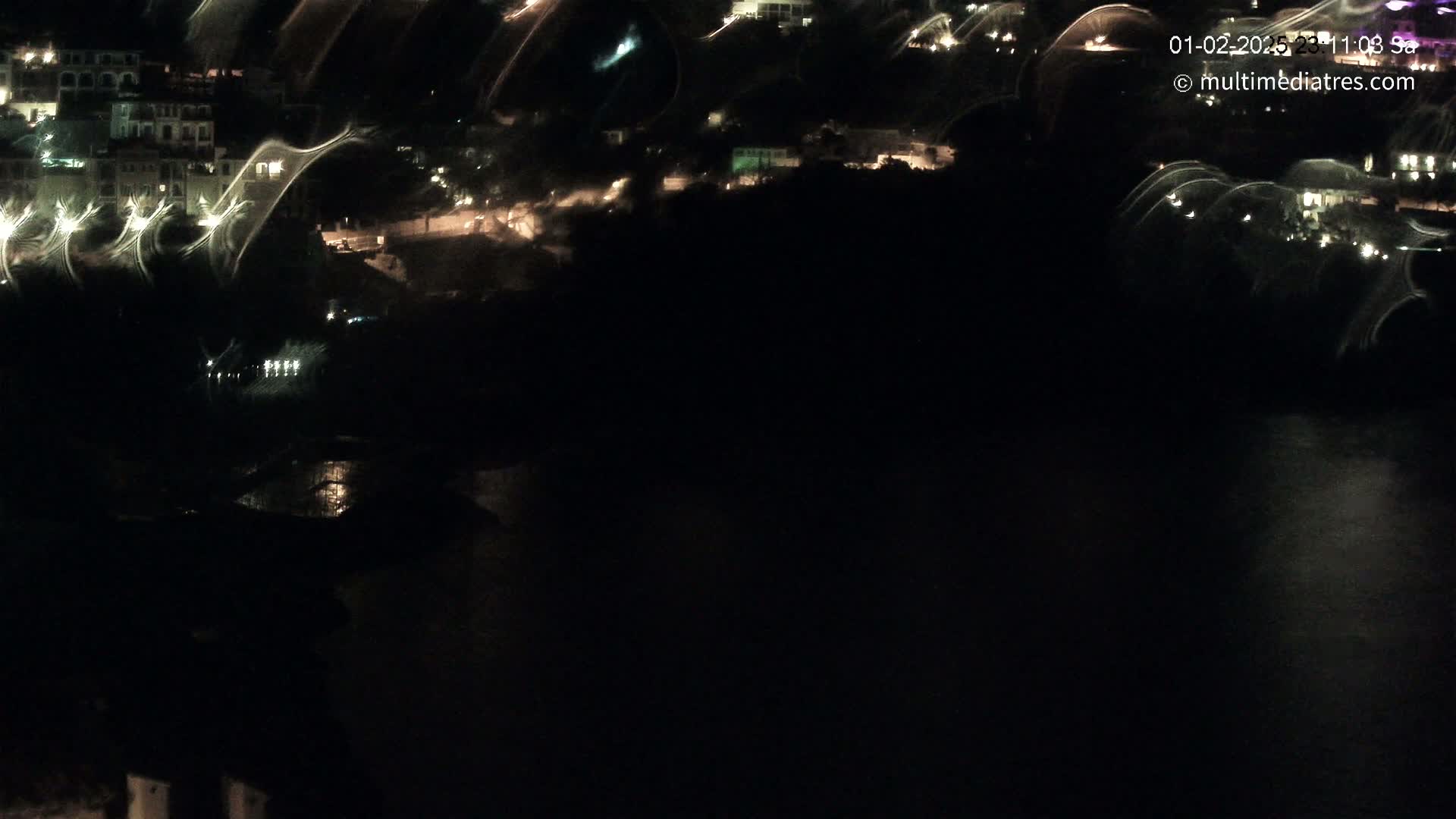 A nighttime, high-angle view shows a dark body of water reflecting the lights of a hillside town.