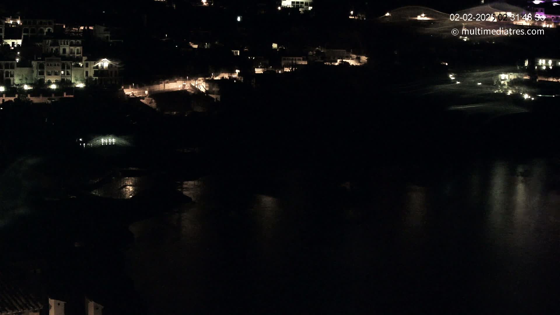 A nighttime, aerial view shows a dark body of water reflecting the lights of a hillside village.