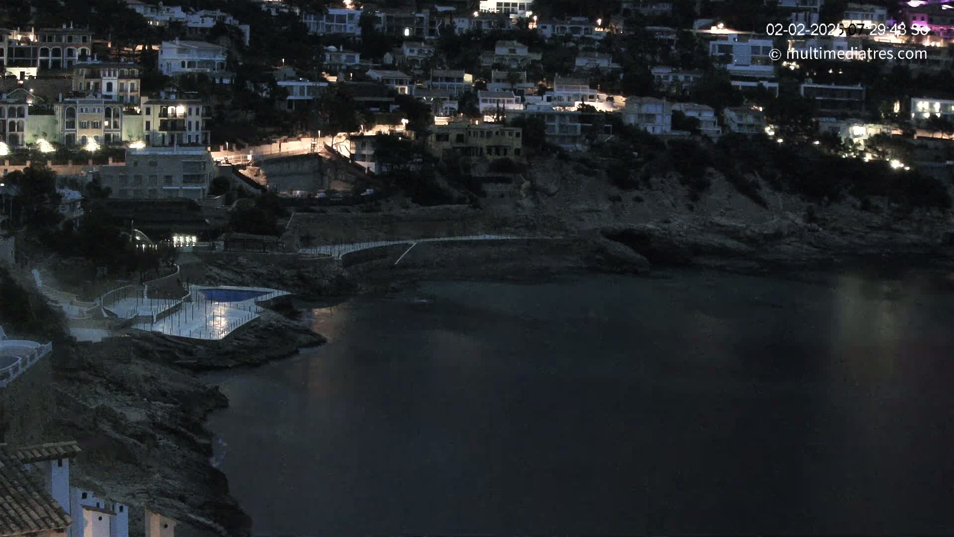 A dark, nighttime view of a coastal town with many buildings lit up, a small pool, and rocky shoreline extending into a calm, dark body of water.