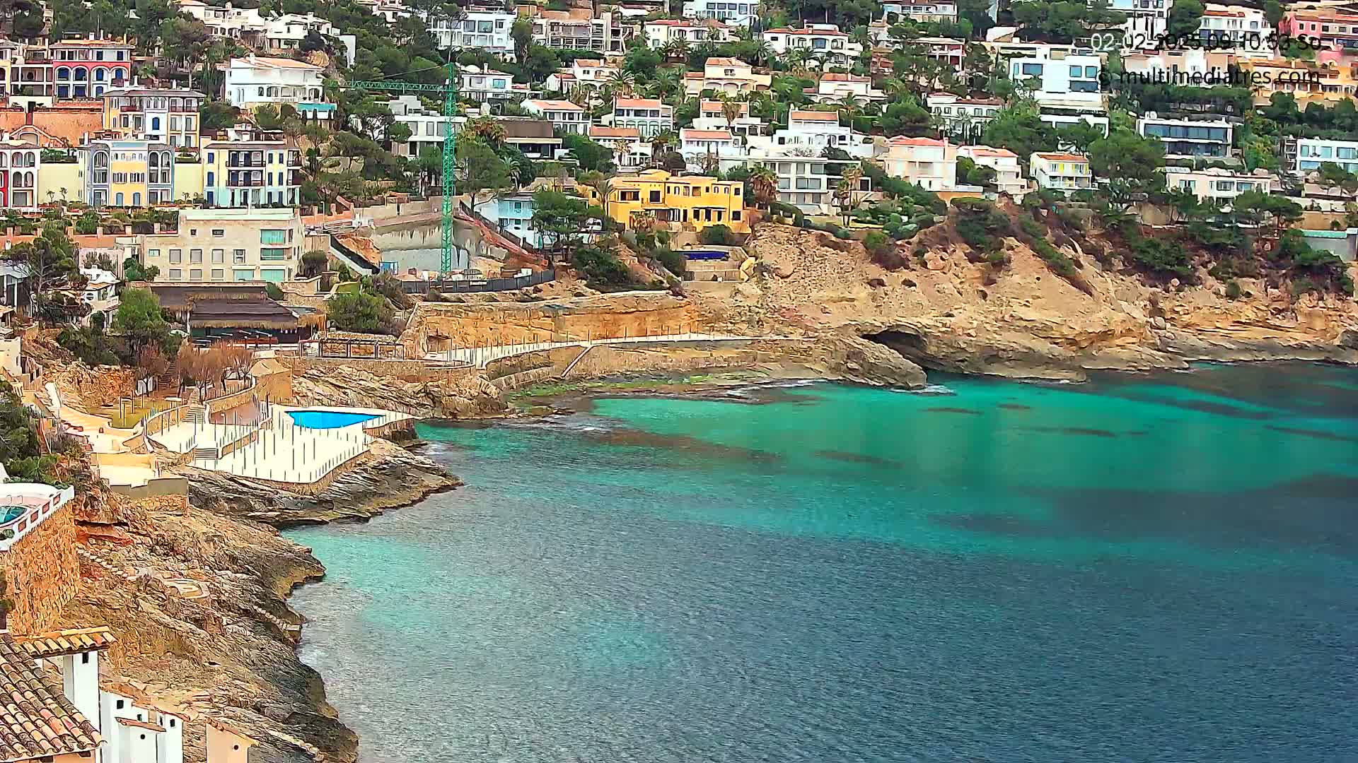 A coastal town with colorful buildings is situated on a rocky hillside overlooking a calm, teal sea with a small pool next to the water.