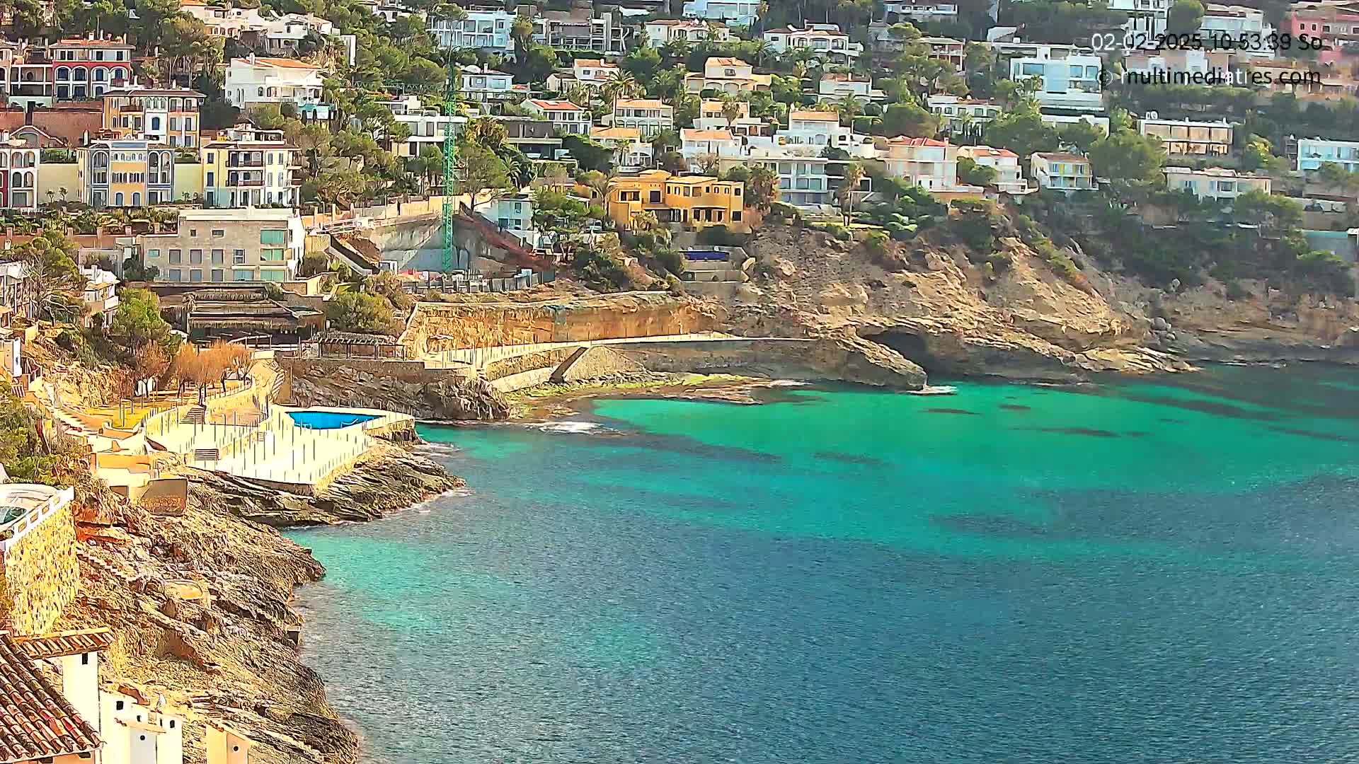 A coastal town with white buildings clings to a rocky hillside overlooking a bright teal sea on a sunny day.