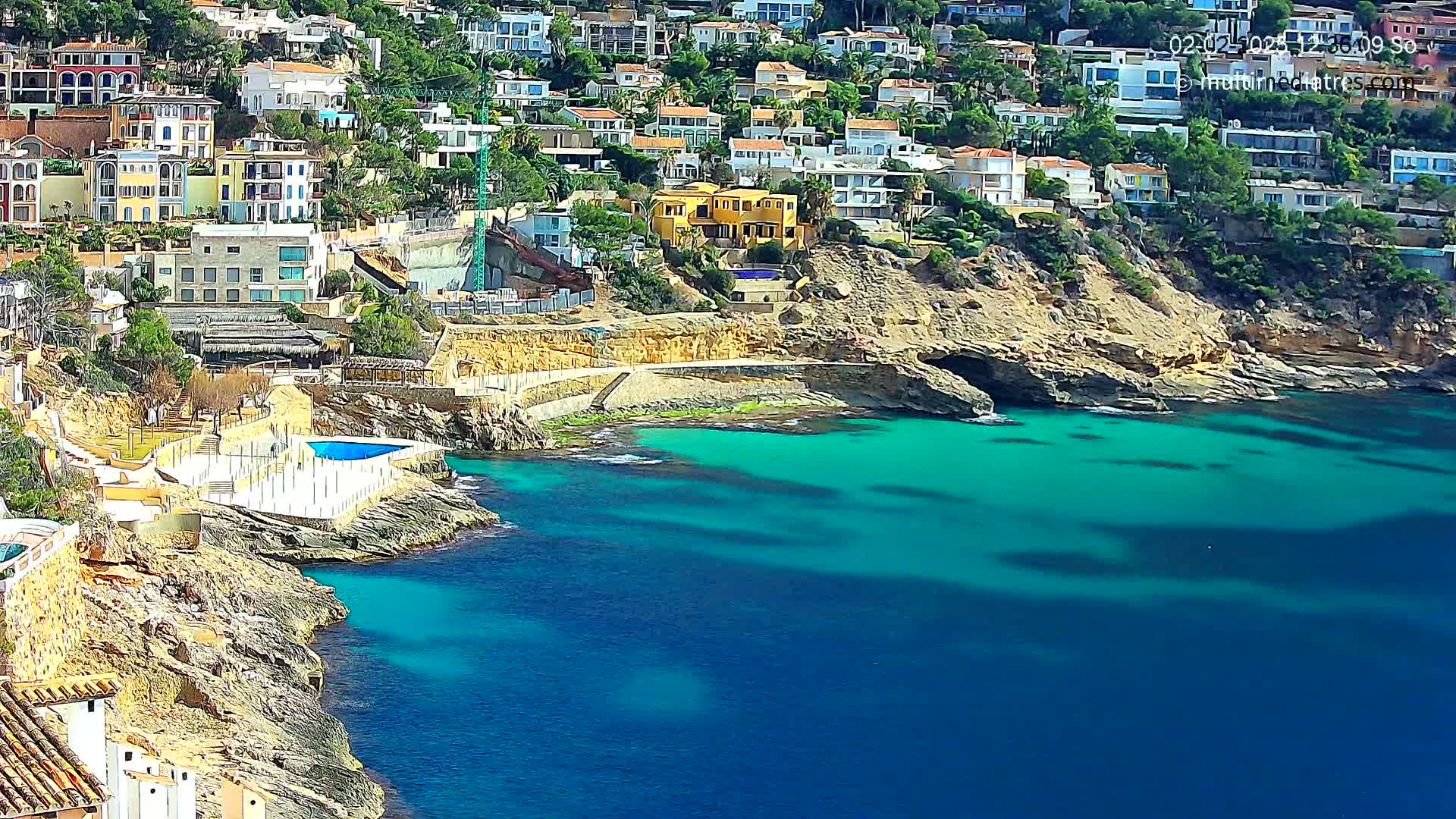 A coastal town with colorful houses built into a rocky hillside overlooking a calm, turquoise sea on a sunny day.
