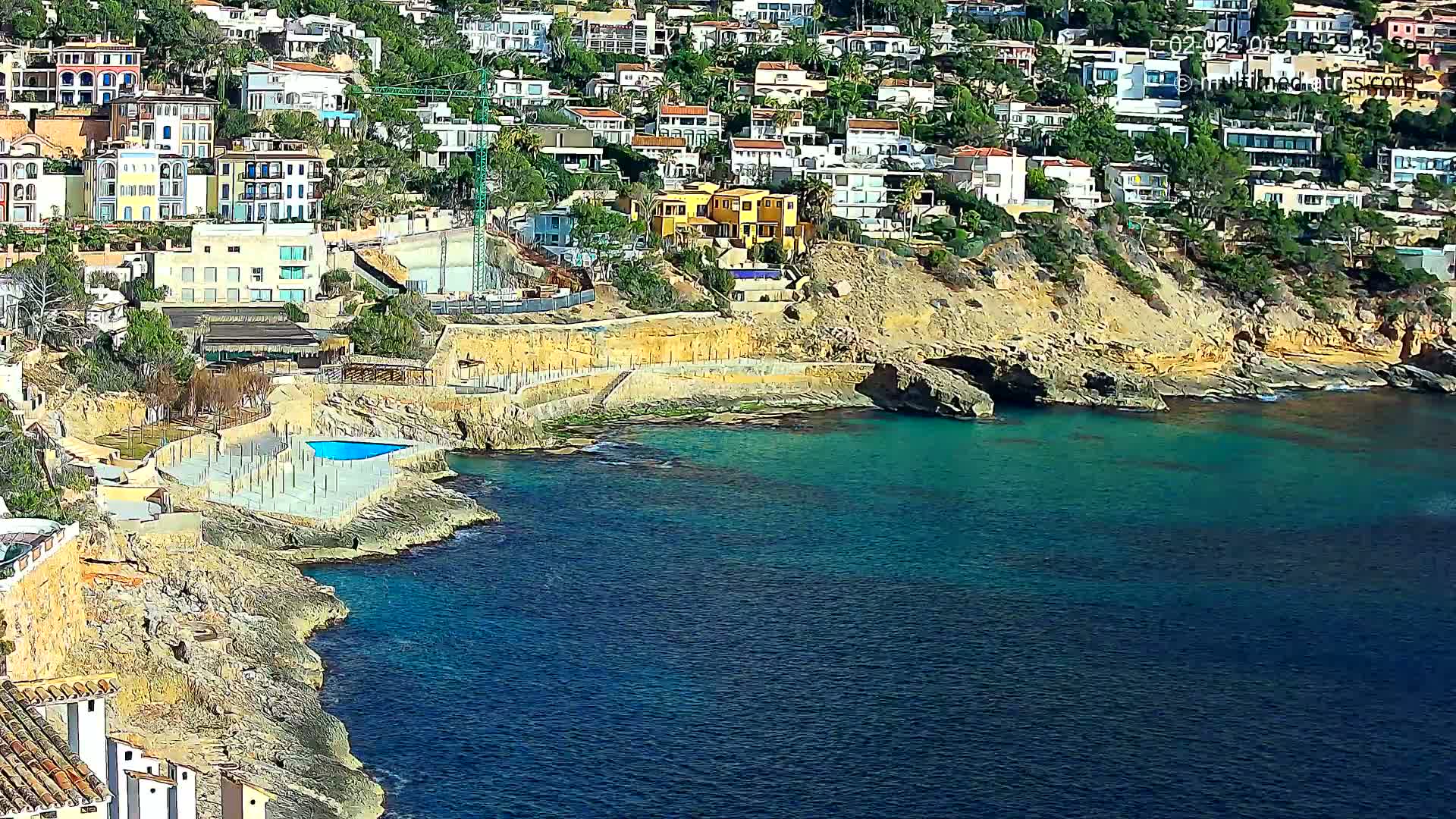 A rocky coastline with teal water features numerous houses built into a hillside, and a small pool is visible near the water's edge.