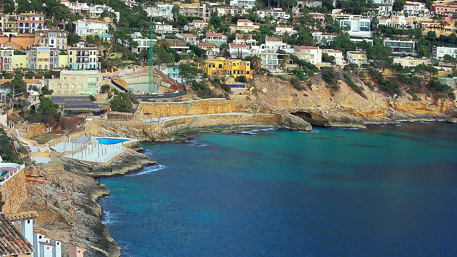 A hillside of houses and buildings overlooks a rocky cove with teal water, a swimming pool, and a small beach area.