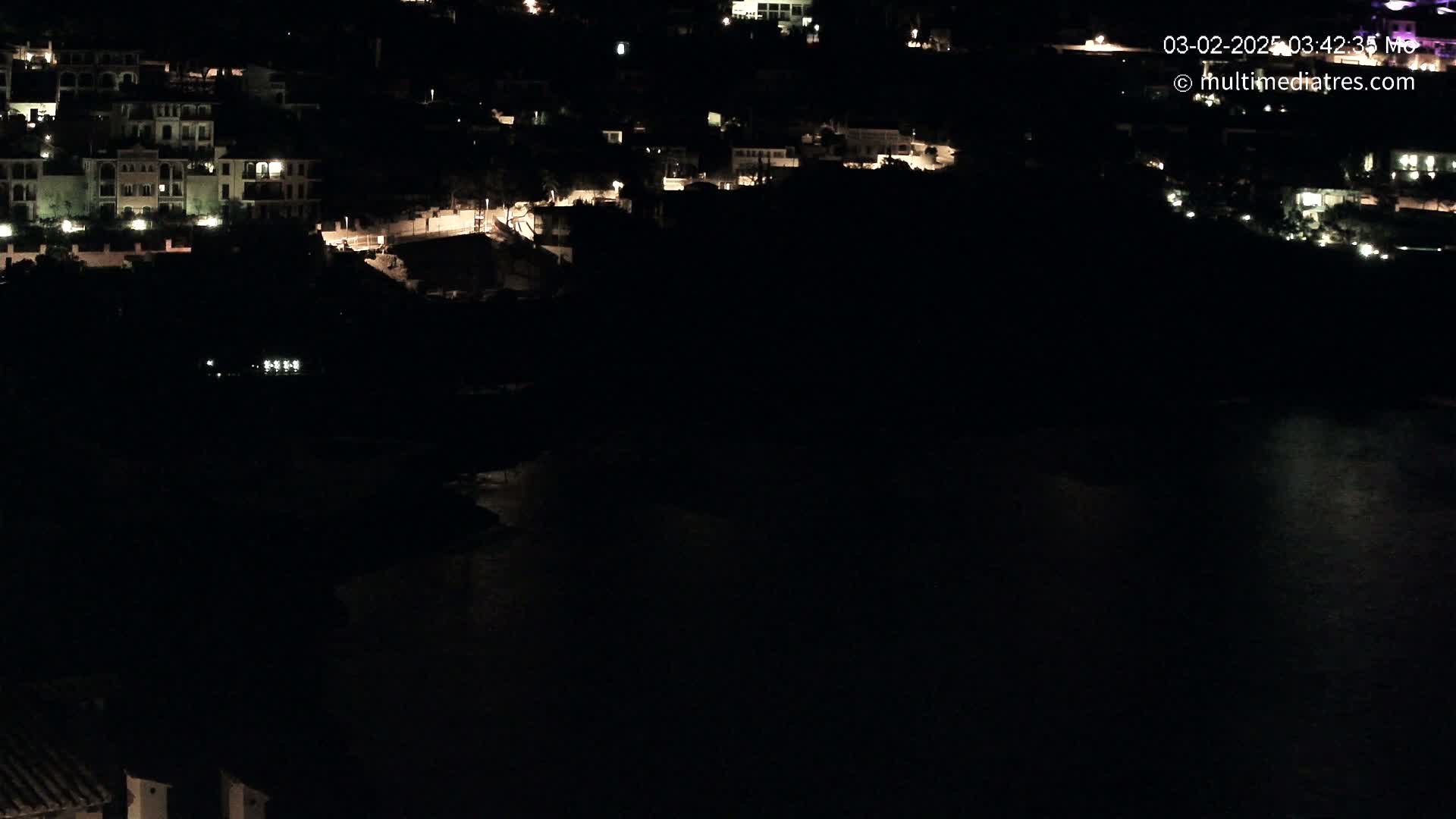 A nighttime, high-angle view shows a dark body of water below a hillside of dimly lit buildings.