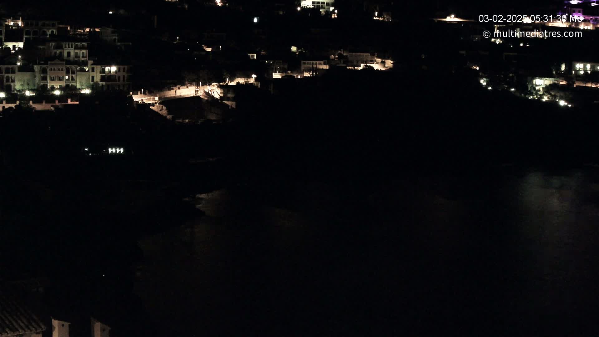 A nighttime, aerial view shows a dark body of water next to a hillside village with dimly lit buildings.