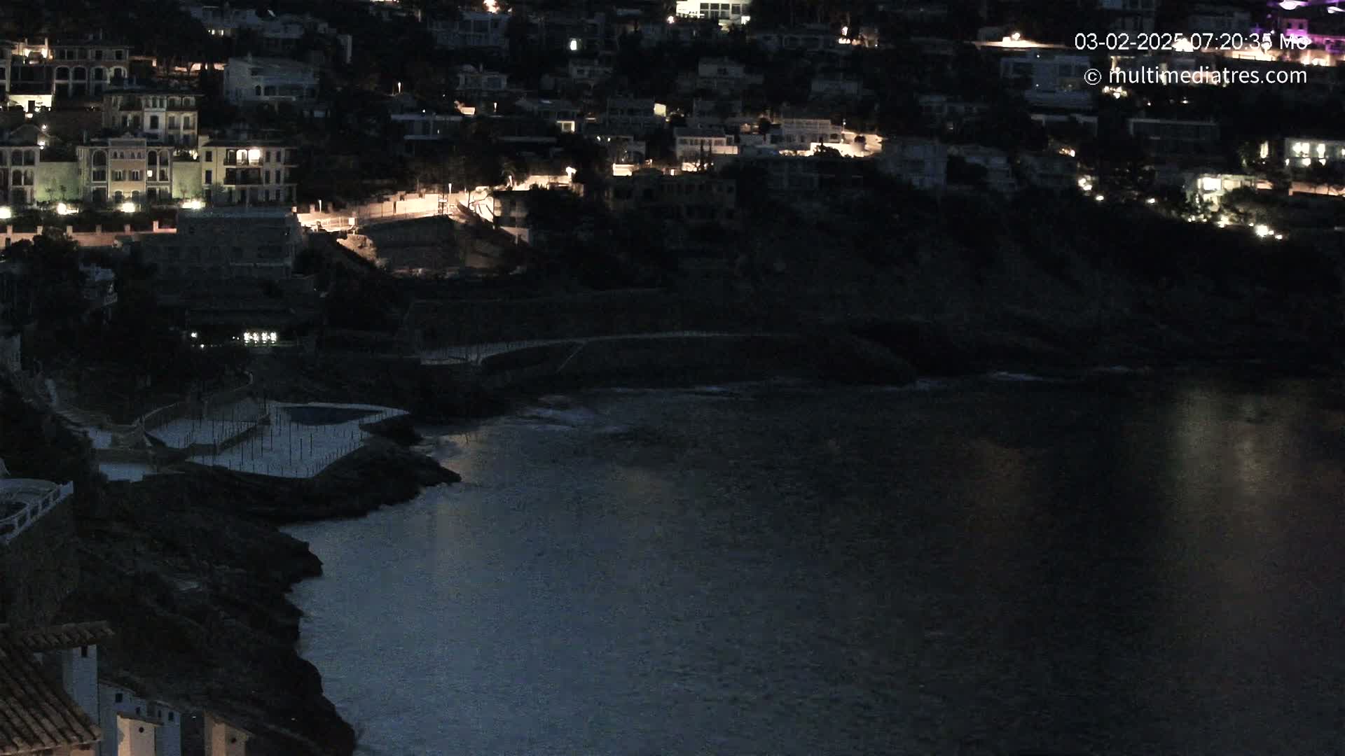 A nighttime view of a dark, calm body of water next to a hillside of buildings with some lights on.