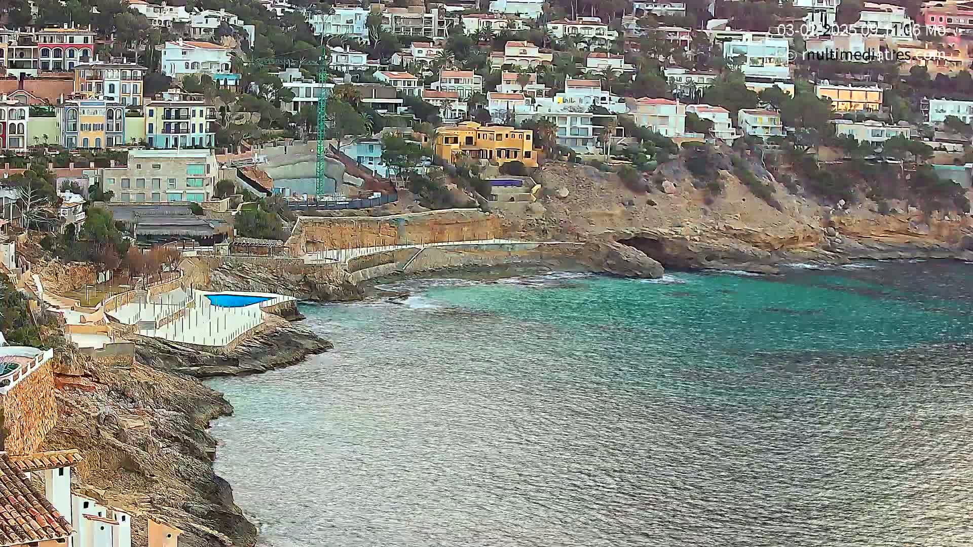 A rocky coastline with a small swimming pool overlooks a calm, turquoise sea, with a hillside of houses behind.
