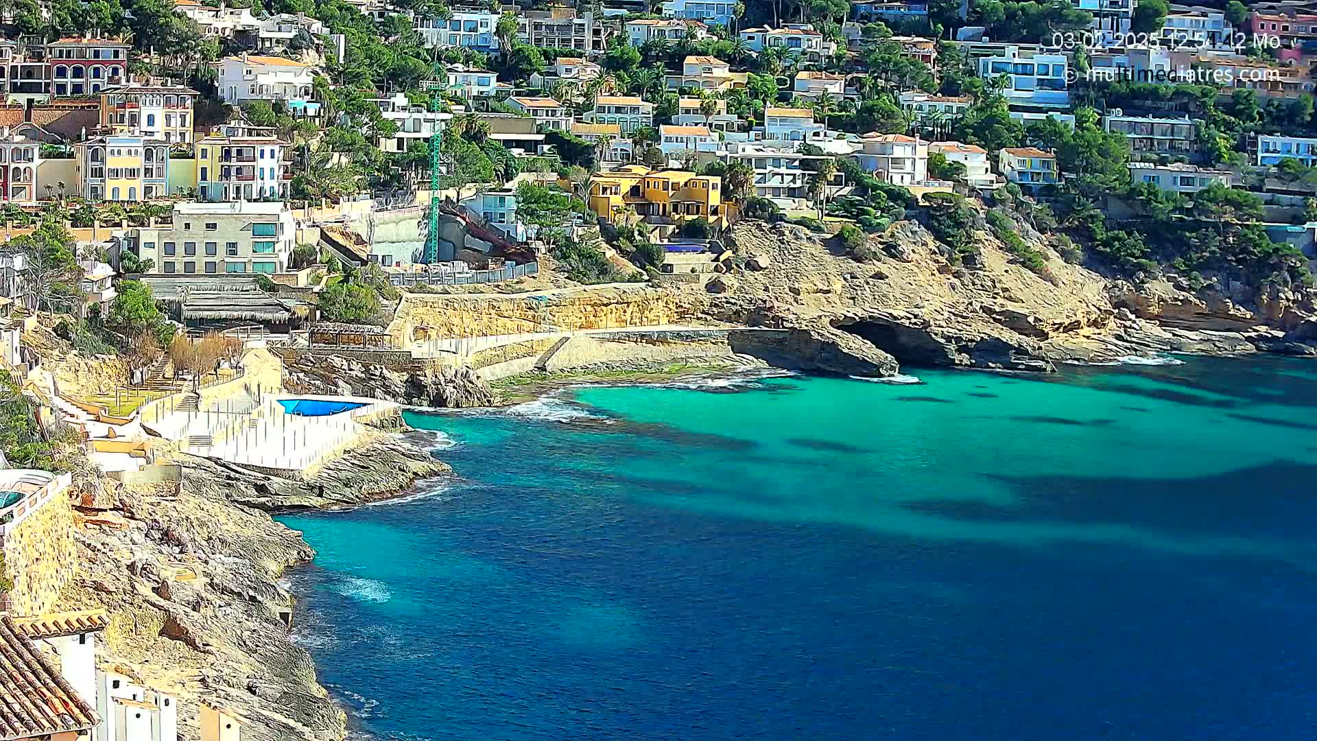 A rocky coastline with turquoise water features many houses built into a hillside on a sunny day.