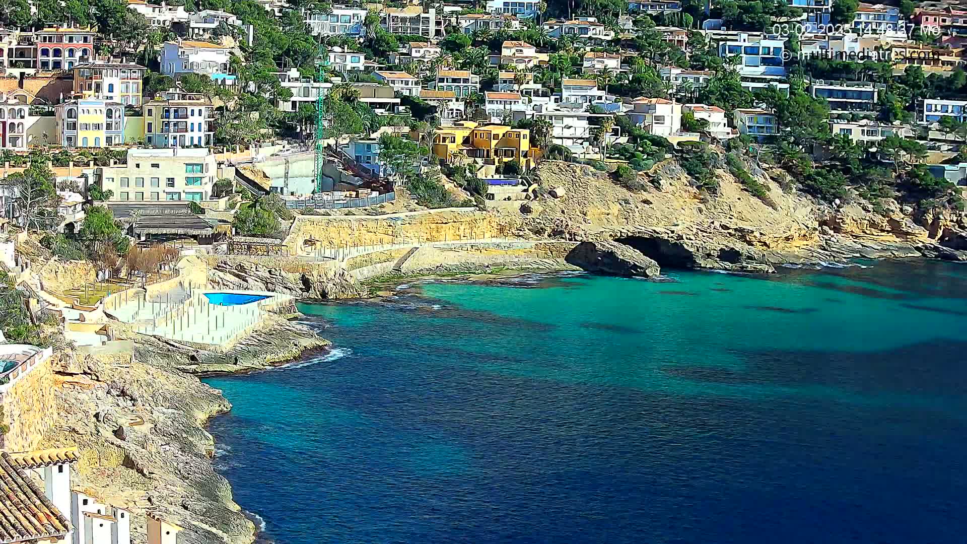 A rocky coast with turquoise water features numerous houses built into a hillside, and a small swimming pool is visible near the water's edge.
