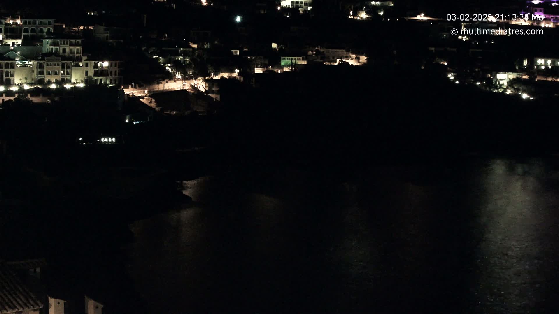 A nighttime view shows a collection of illuminated buildings near a dark body of water.