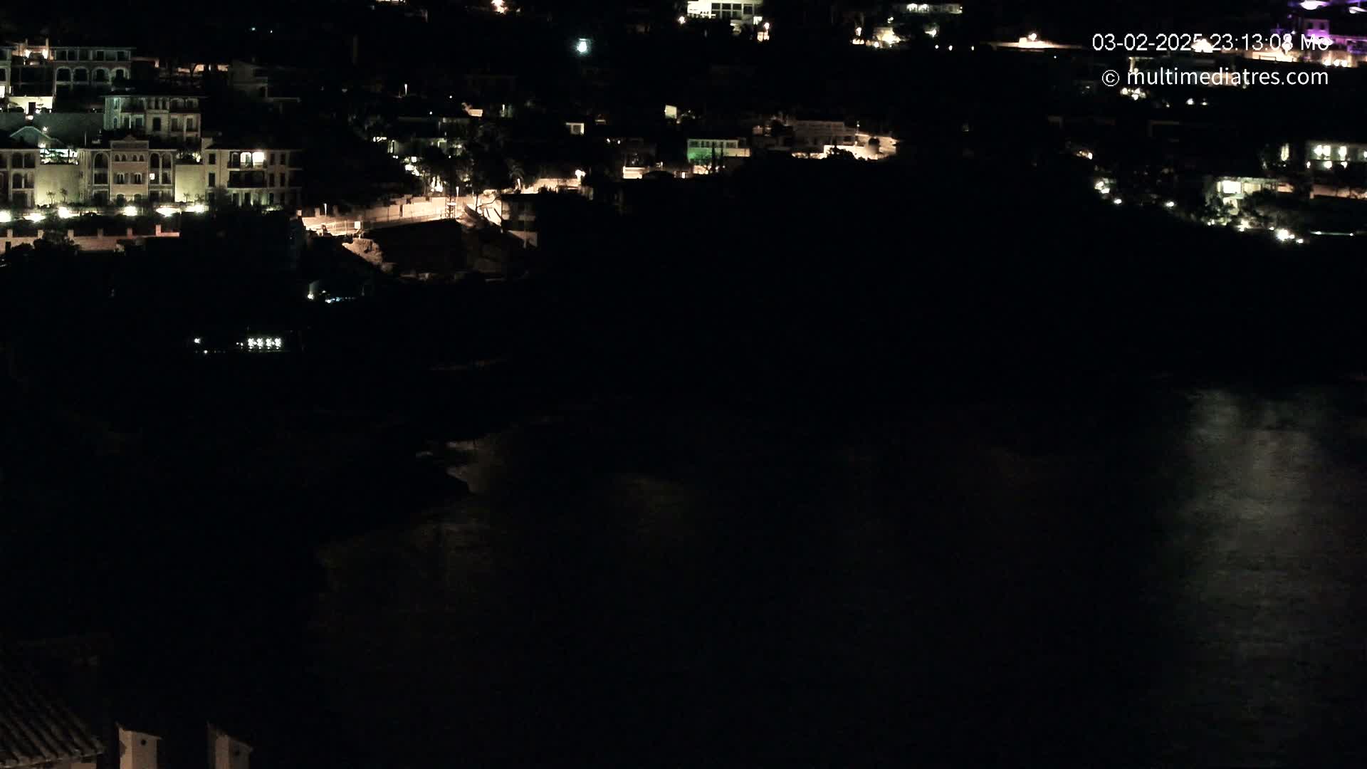 A nighttime aerial view shows a coastal town with illuminated buildings overlooking a dark body of water.