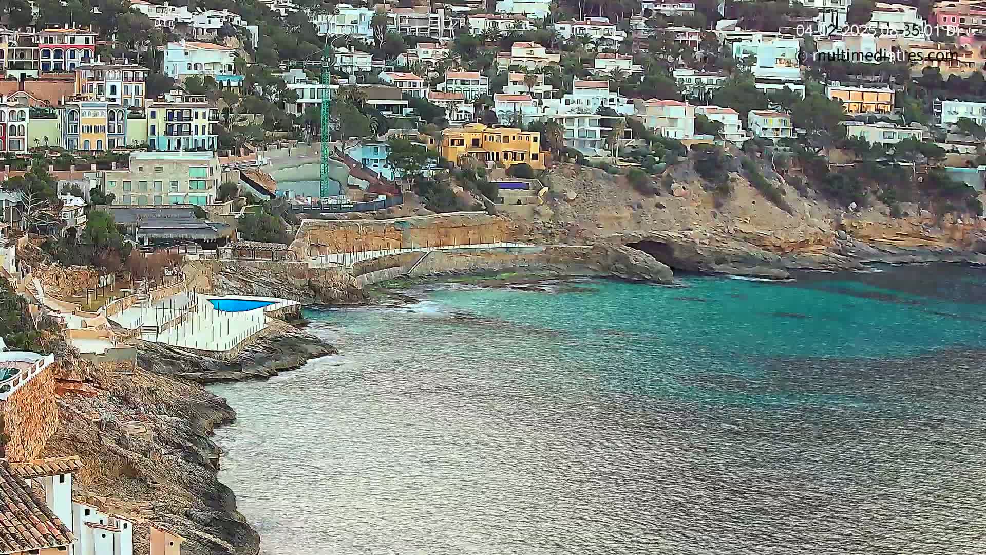 A coastal town built on a rocky hillside overlooks a calm, teal-colored sea with a small, sheltered cove containing a swimming pool.