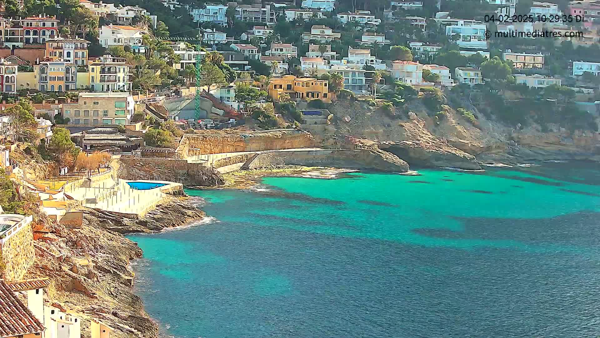 A sunny day reveals a rocky coastline with teal water, a small pool, and a hillside of houses.