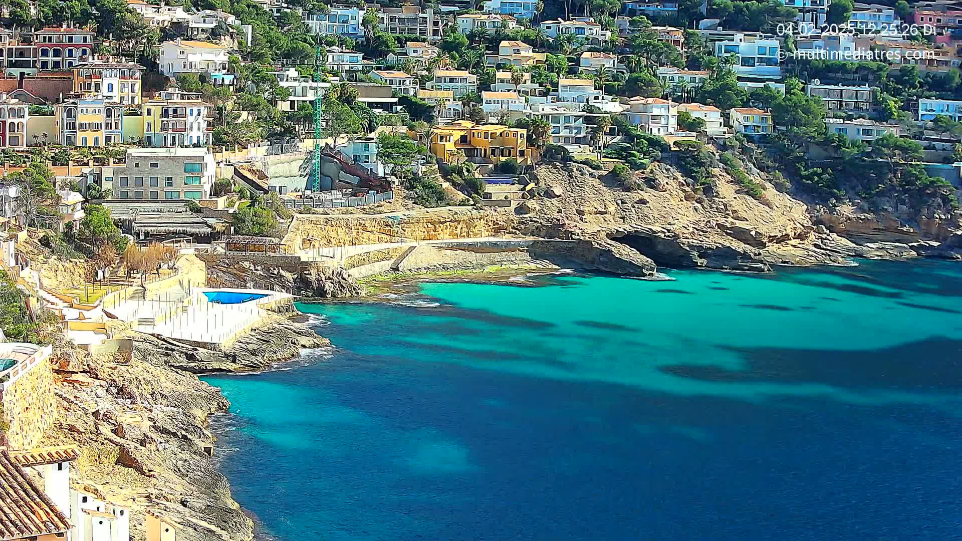 A rocky coastline with turquoise water, featuring a small beach and numerous houses built into a hillside on a sunny day.