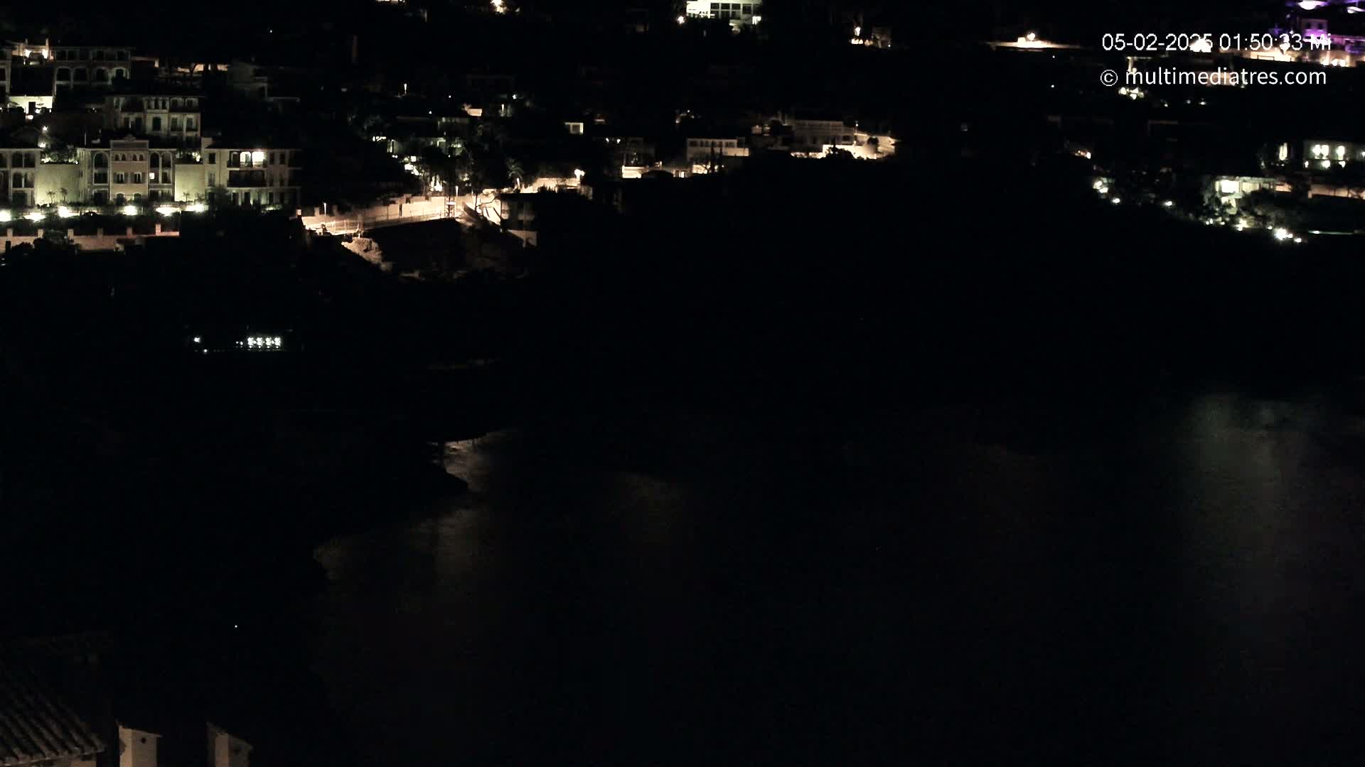 A nighttime, aerial view shows a dark body of water reflecting dim light, beside a hillside of dimly lit buildings.