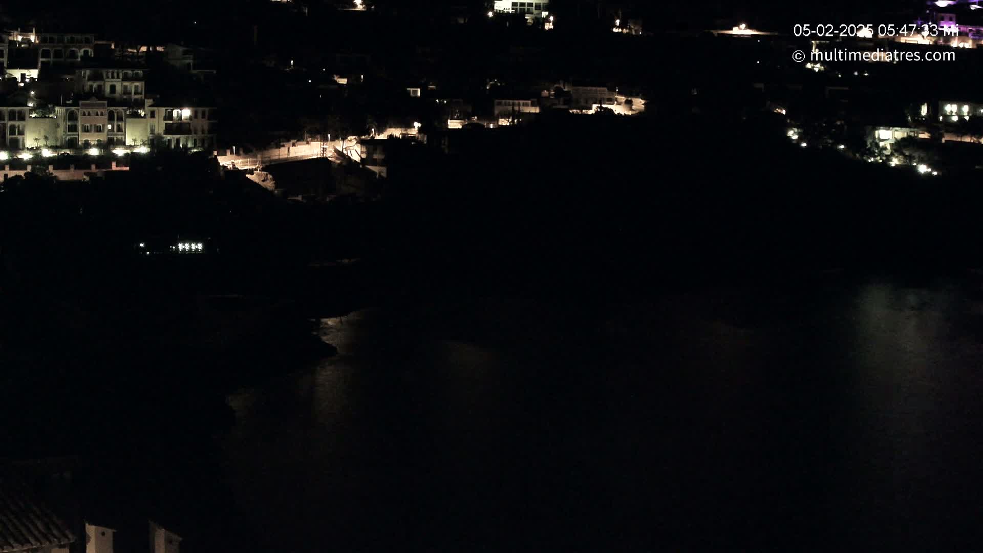 A nighttime aerial view shows a collection of illuminated buildings near a dark body of water.