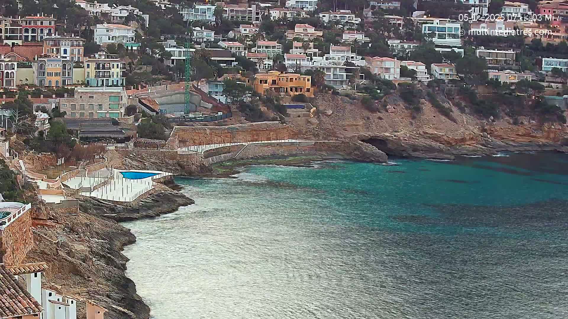 A rocky coastline with turquoise water features a cluster of buildings perched on a hillside, a small pool, and a paved walkway.