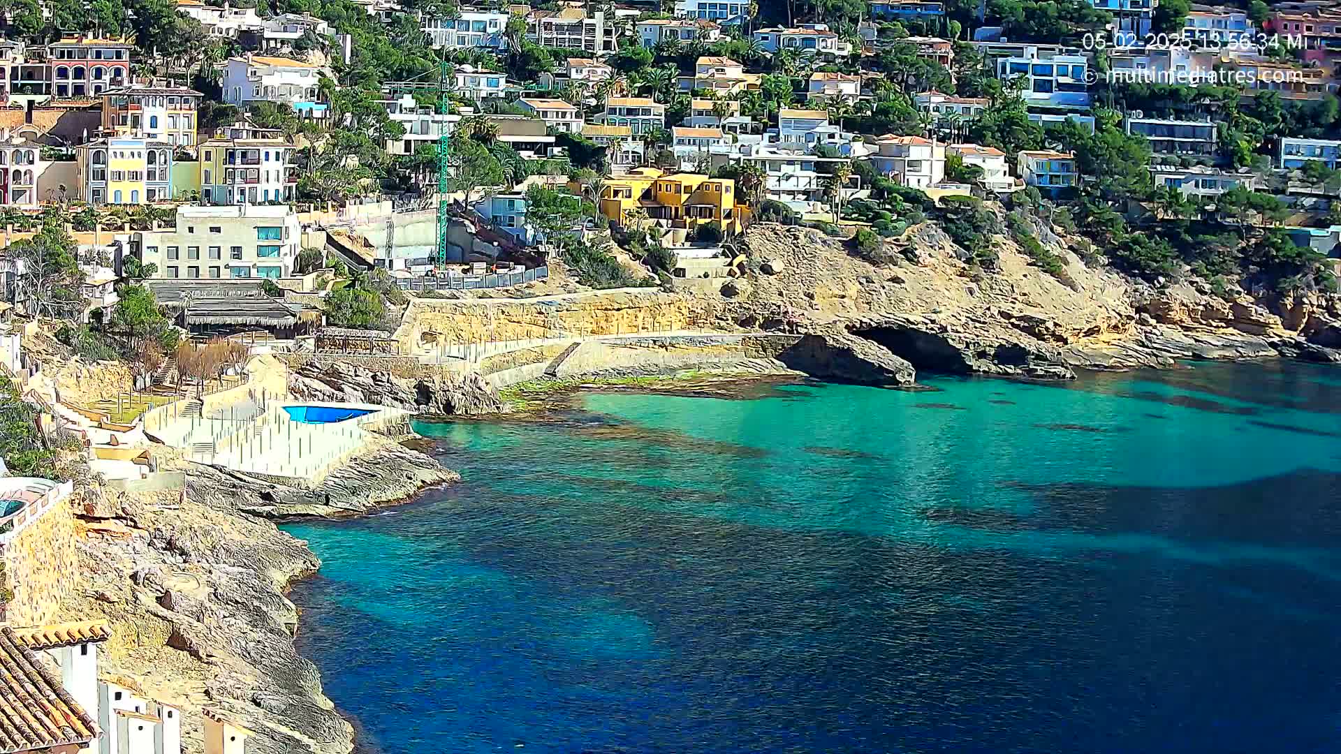A rocky coastline with turquoise water, featuring a collection of buildings on a hillside, and a small swimming pool near the water's edge under sunny skies.