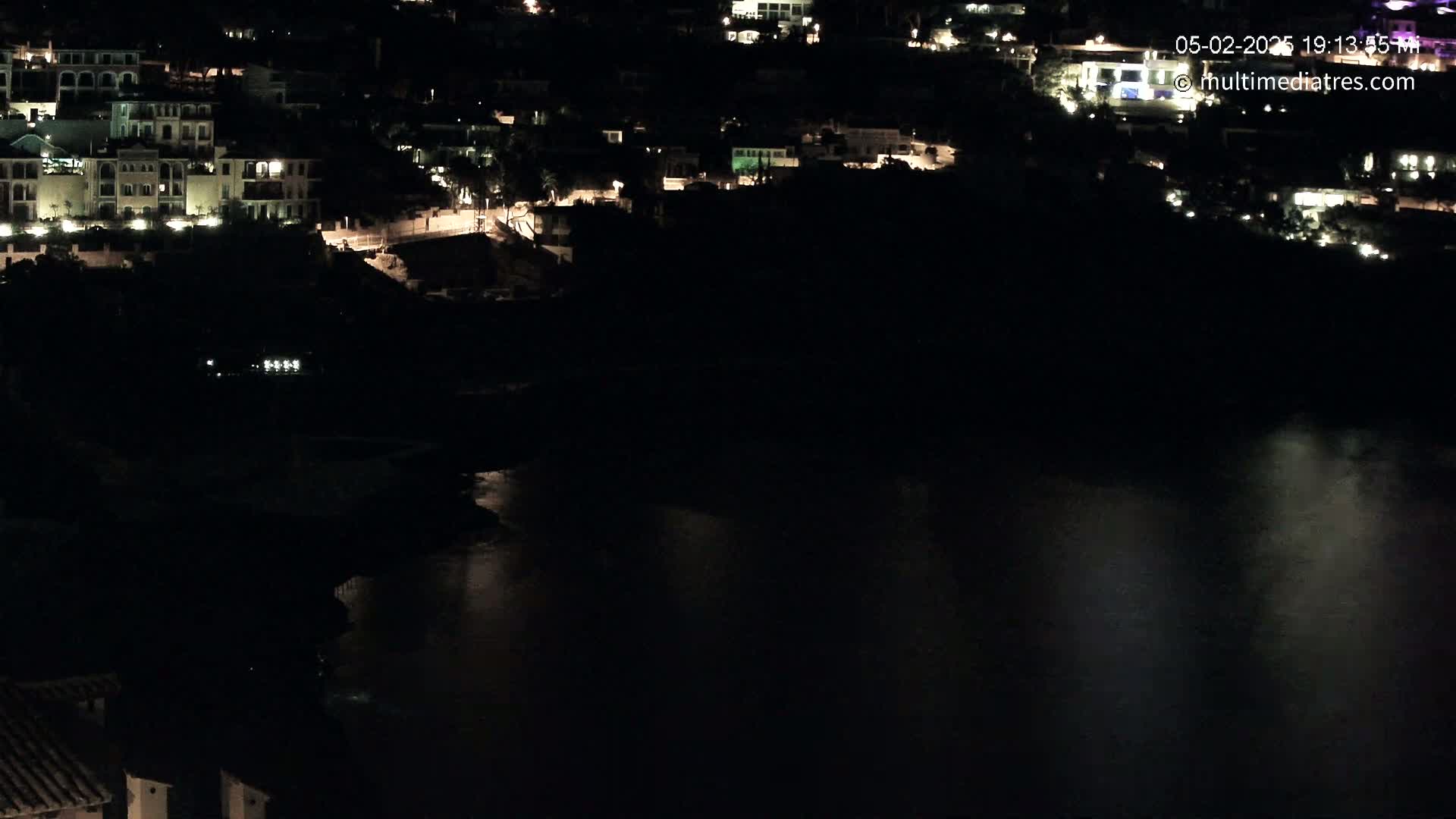 A nighttime view of a waterfront town with lights illuminating buildings and the dark water reflecting some of the light.