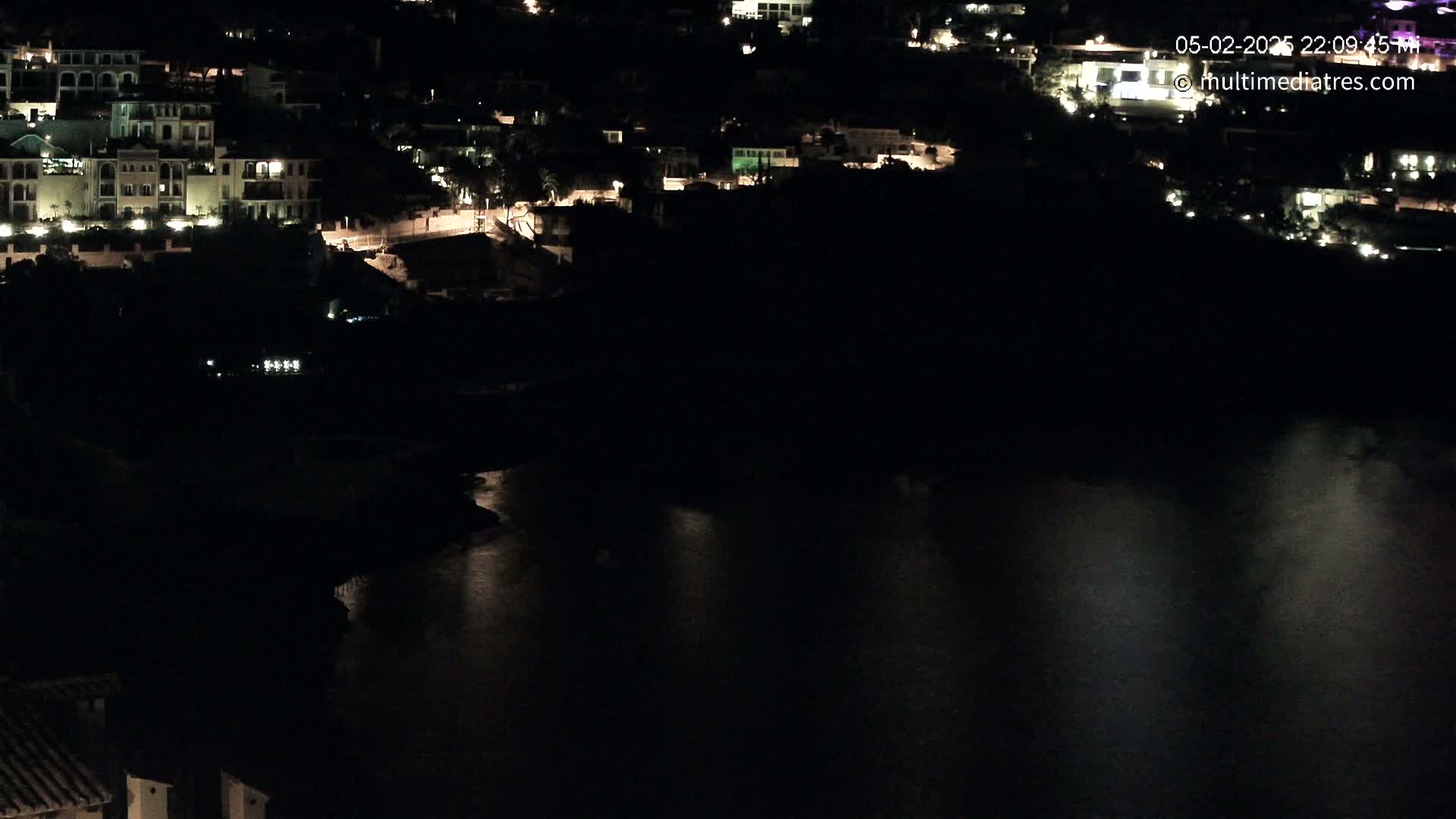 A nighttime view shows a dark body of water reflecting the lights of buildings on a hillside.