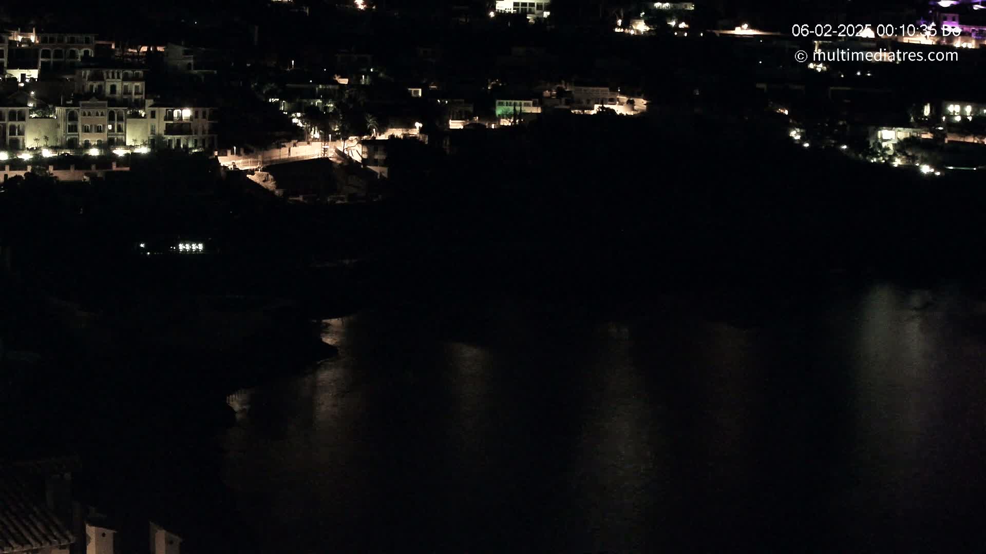 A nighttime, aerial view shows a dark body of water reflecting the lights of buildings on a hillside.