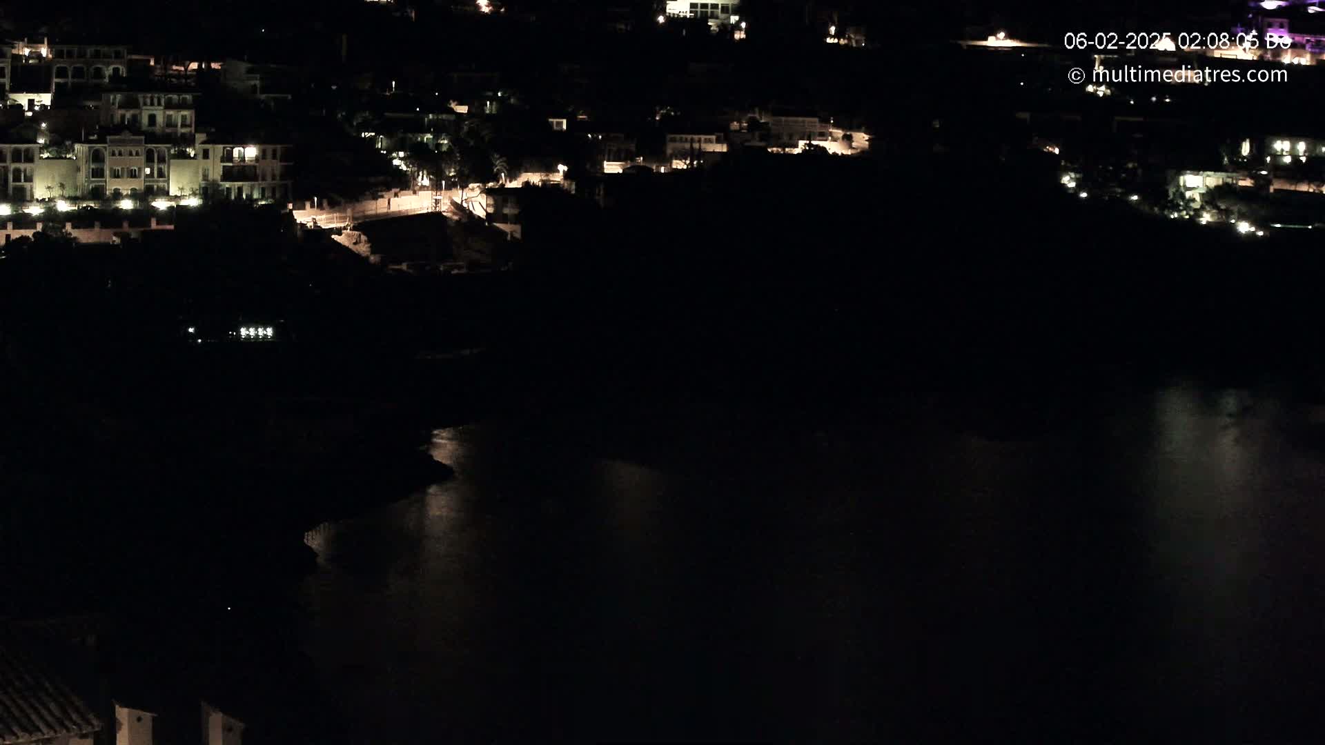 A nighttime view shows a body of dark water reflecting the lights of buildings on a hillside.