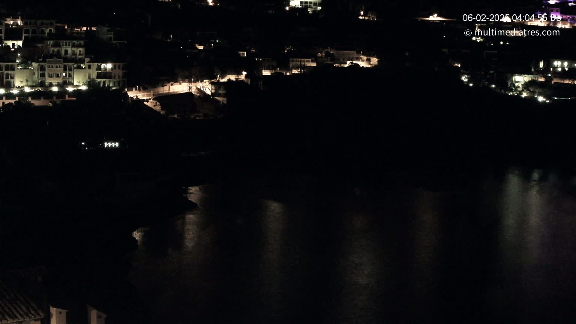 A nighttime view of a coastal town with buildings illuminated against a dark body of water reflecting the lights.