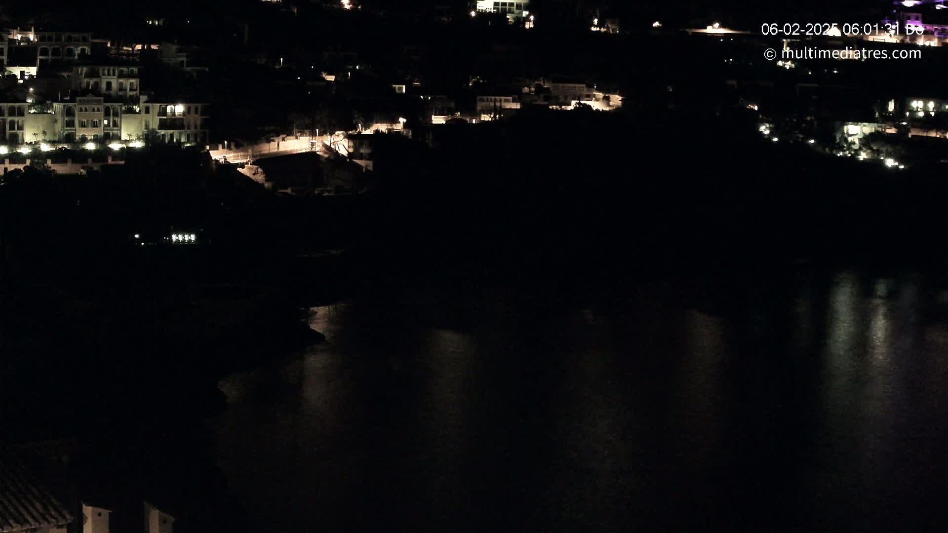 A nighttime view of a waterfront town with illuminated buildings and a dark body of water reflecting some light.