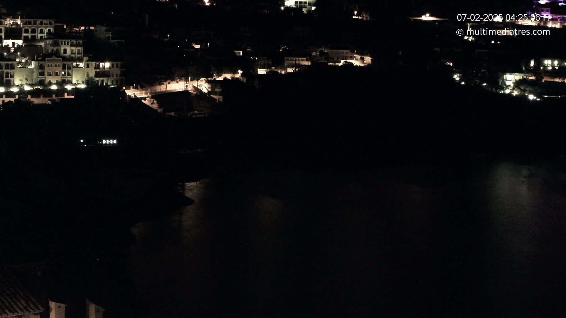 A nighttime, high-angle view shows a dark body of water reflecting the lights from buildings on a hillside.