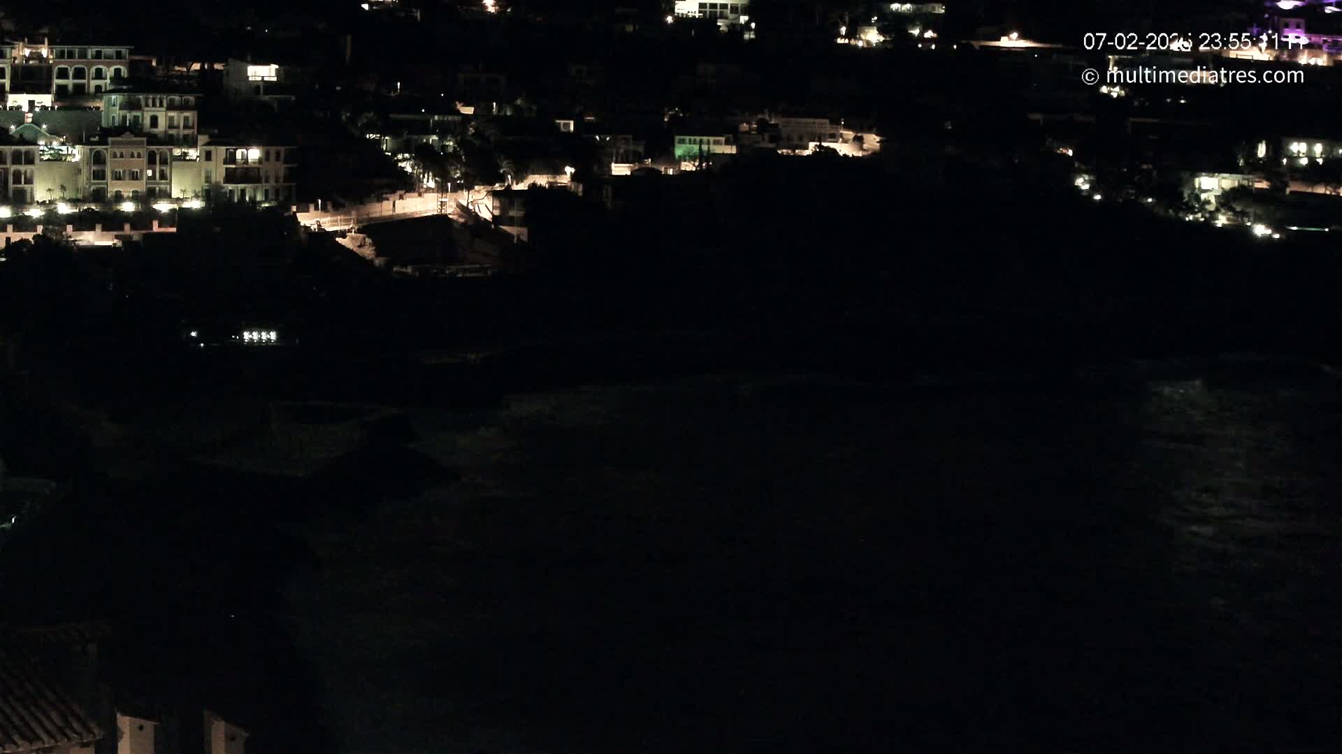 A nighttime coastal scene shows a dark body of water below a hillside of dimly lit buildings.