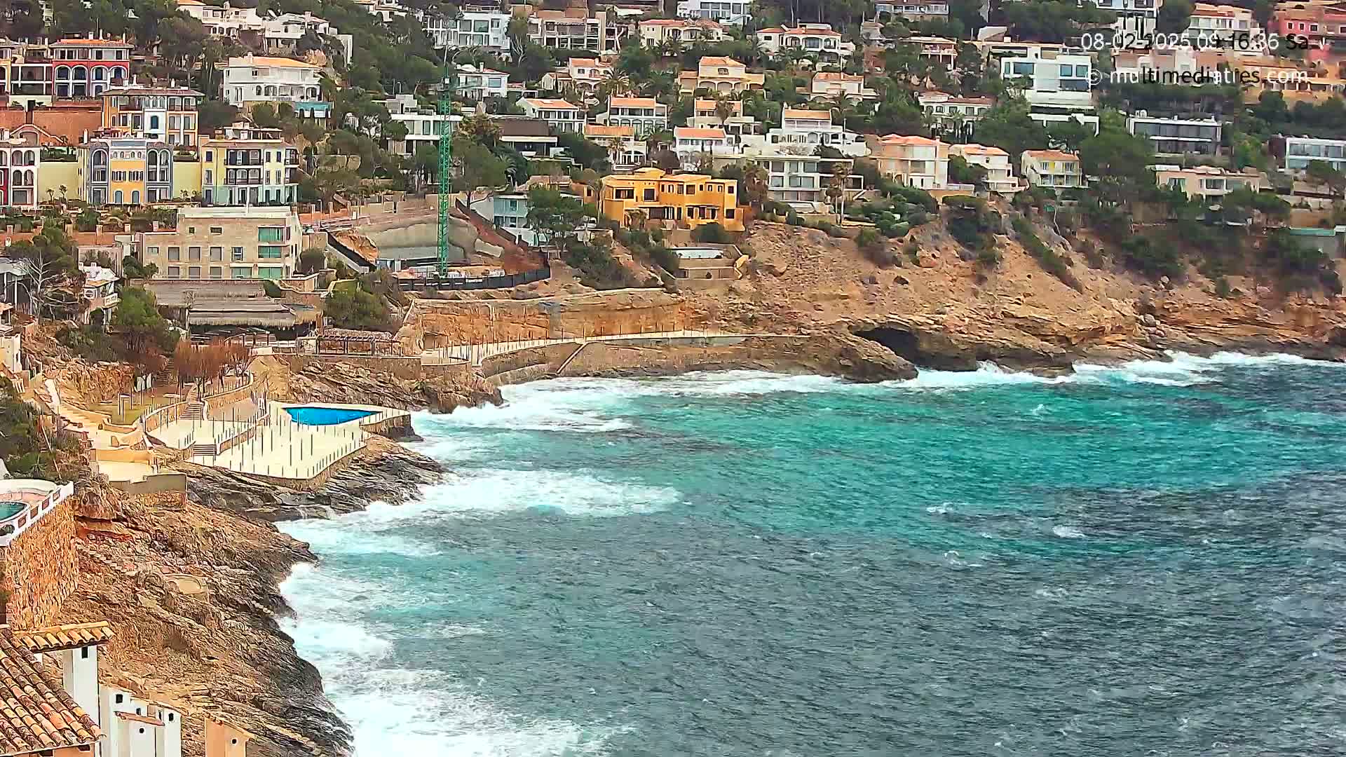 Waves crash against a rocky coastline where numerous houses cling to a hillside overlooking a turquoise sea.