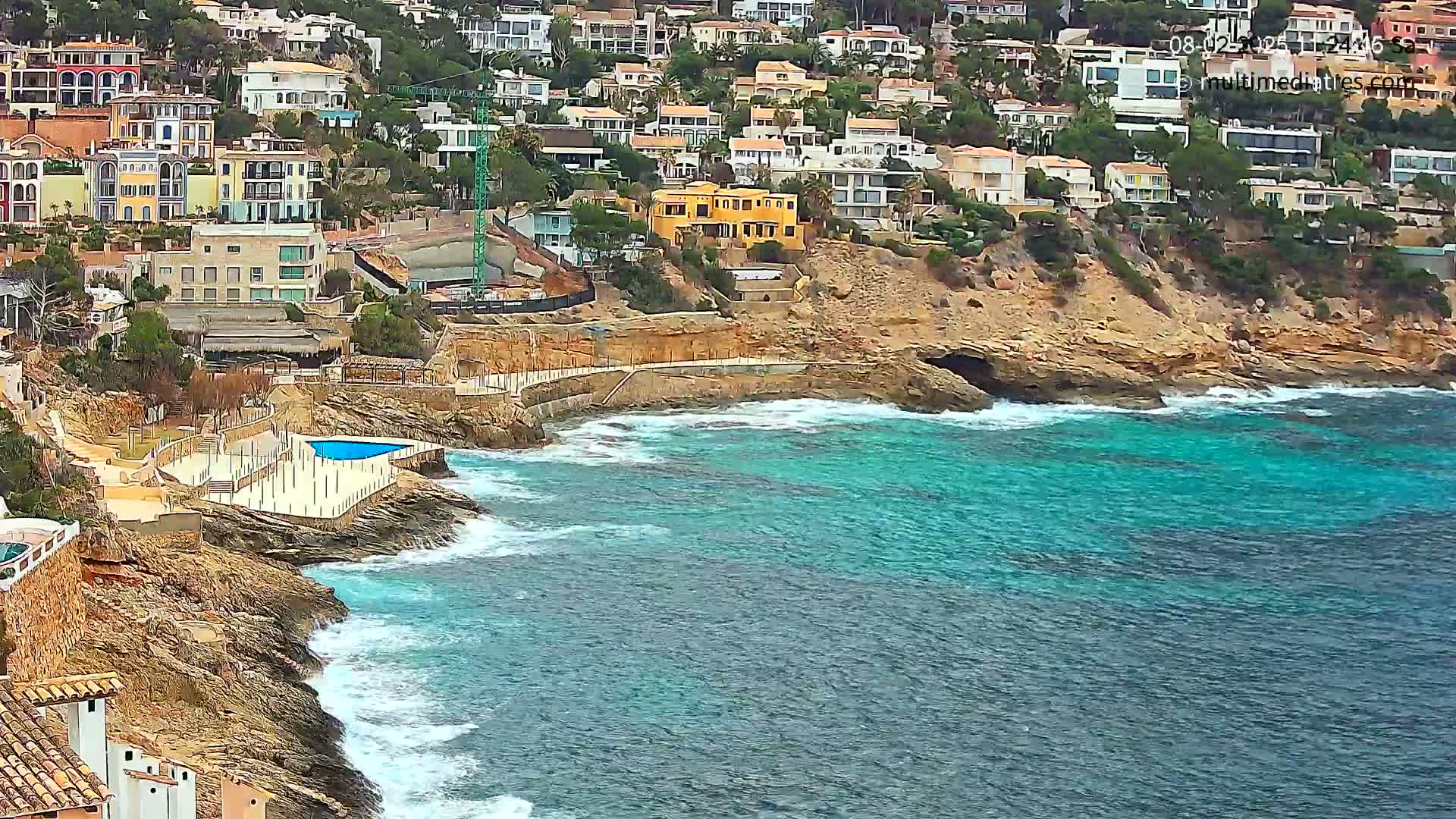 A rocky coastline with turquoise water and white waves features a cluster of buildings, a small swimming pool, and some ongoing construction.
