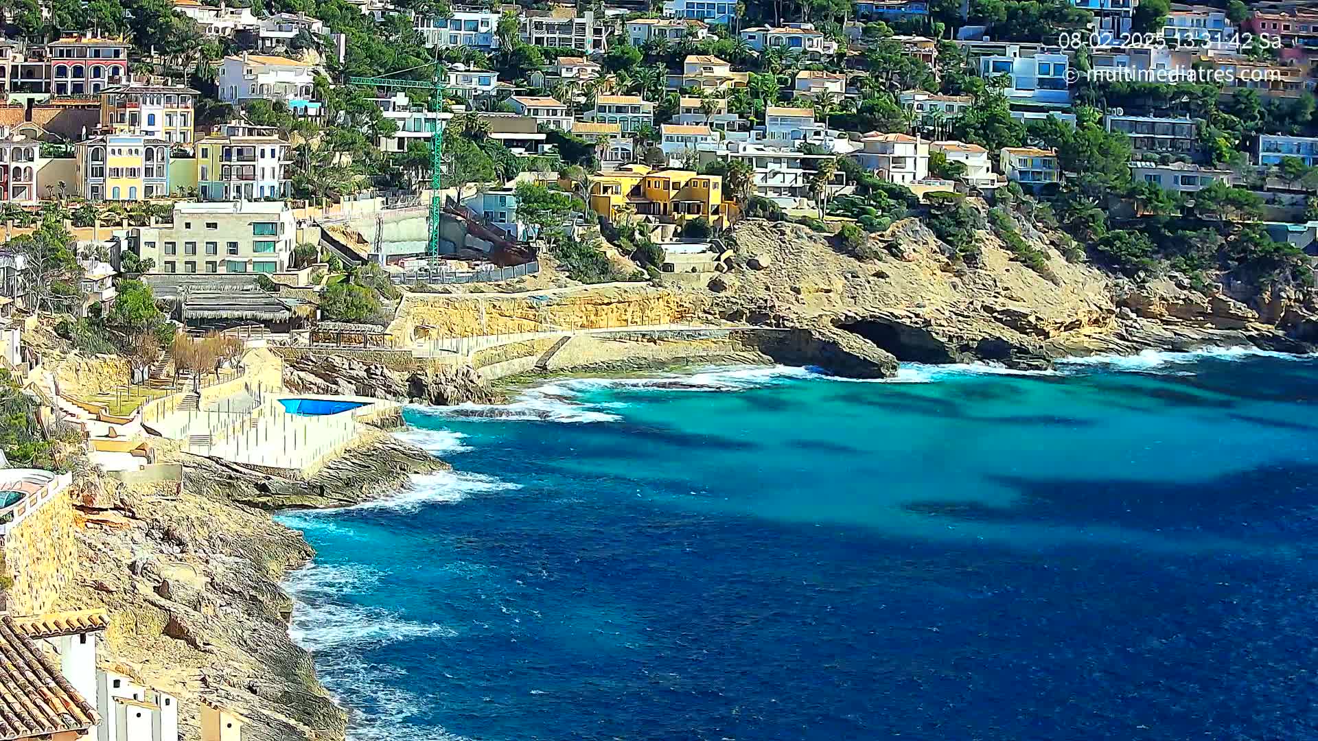 A rocky coastline with numerous houses built into a hillside overlooks a vibrant turquoise ocean with small waves.
