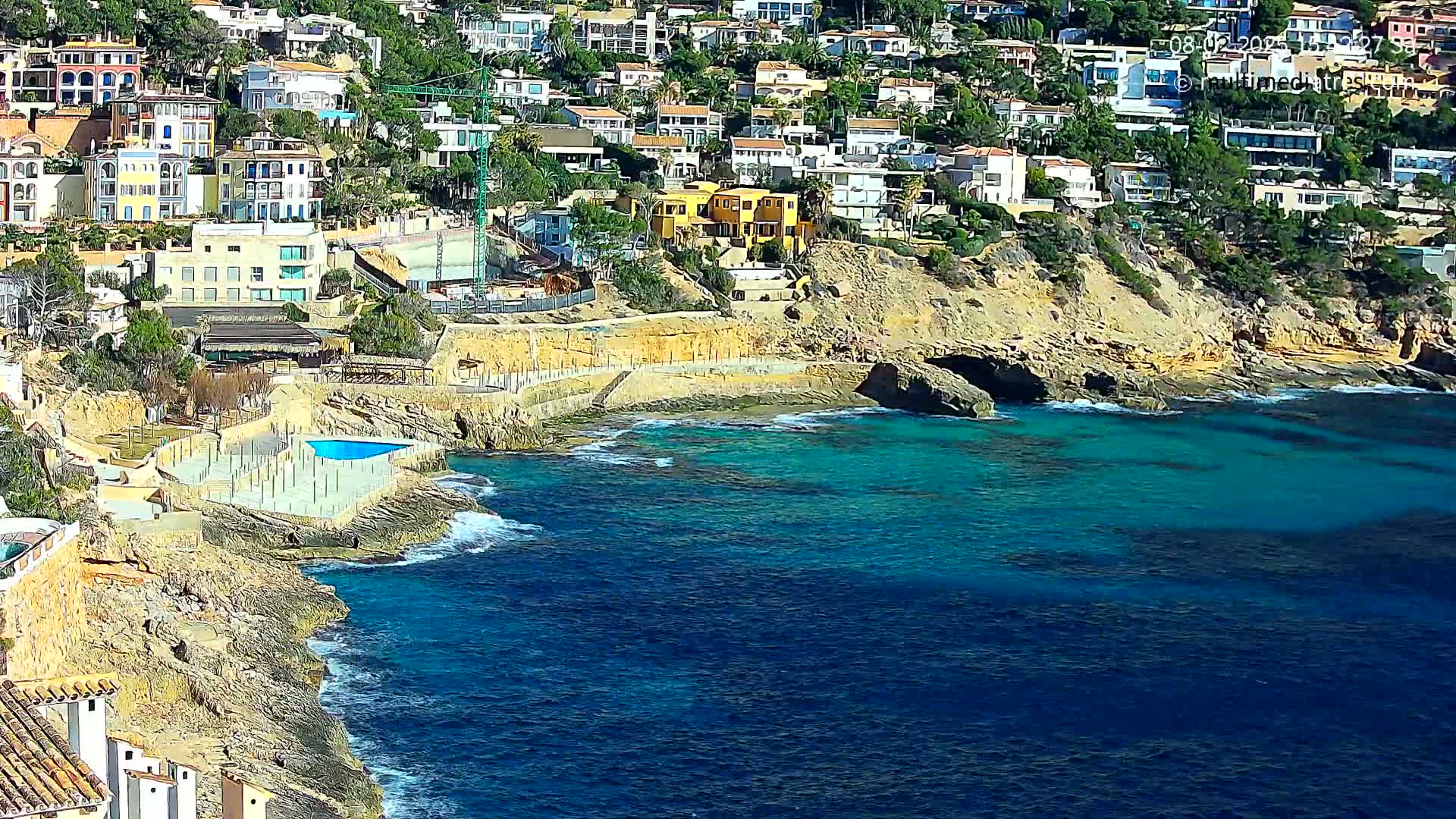 A coastal town with many houses on a hillside overlooks a rocky shoreline and clear turquoise water on a sunny day.